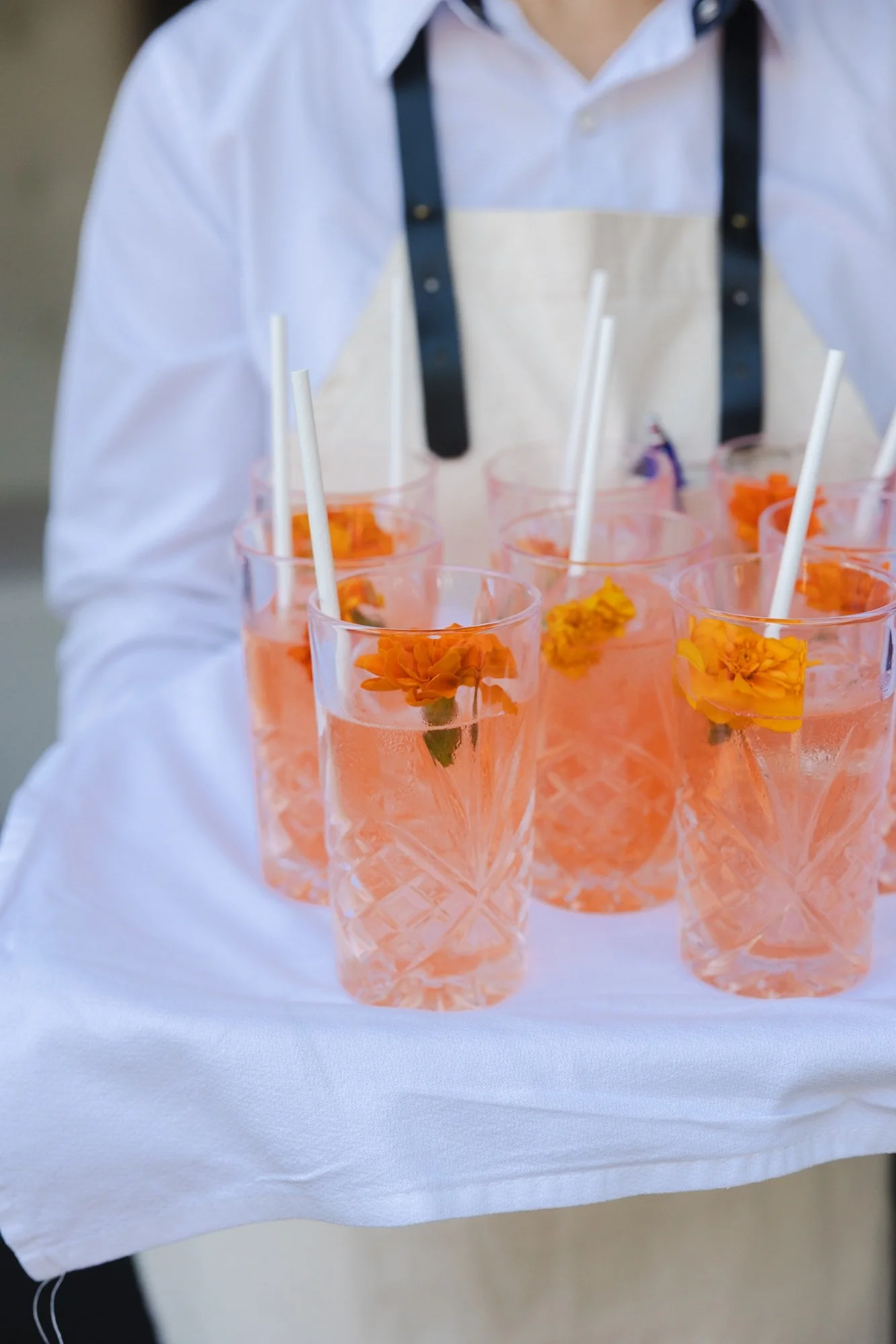 A person holding a white tray with six glasses filled with pink liquid and decorated with orange flowers, each with a white straw.