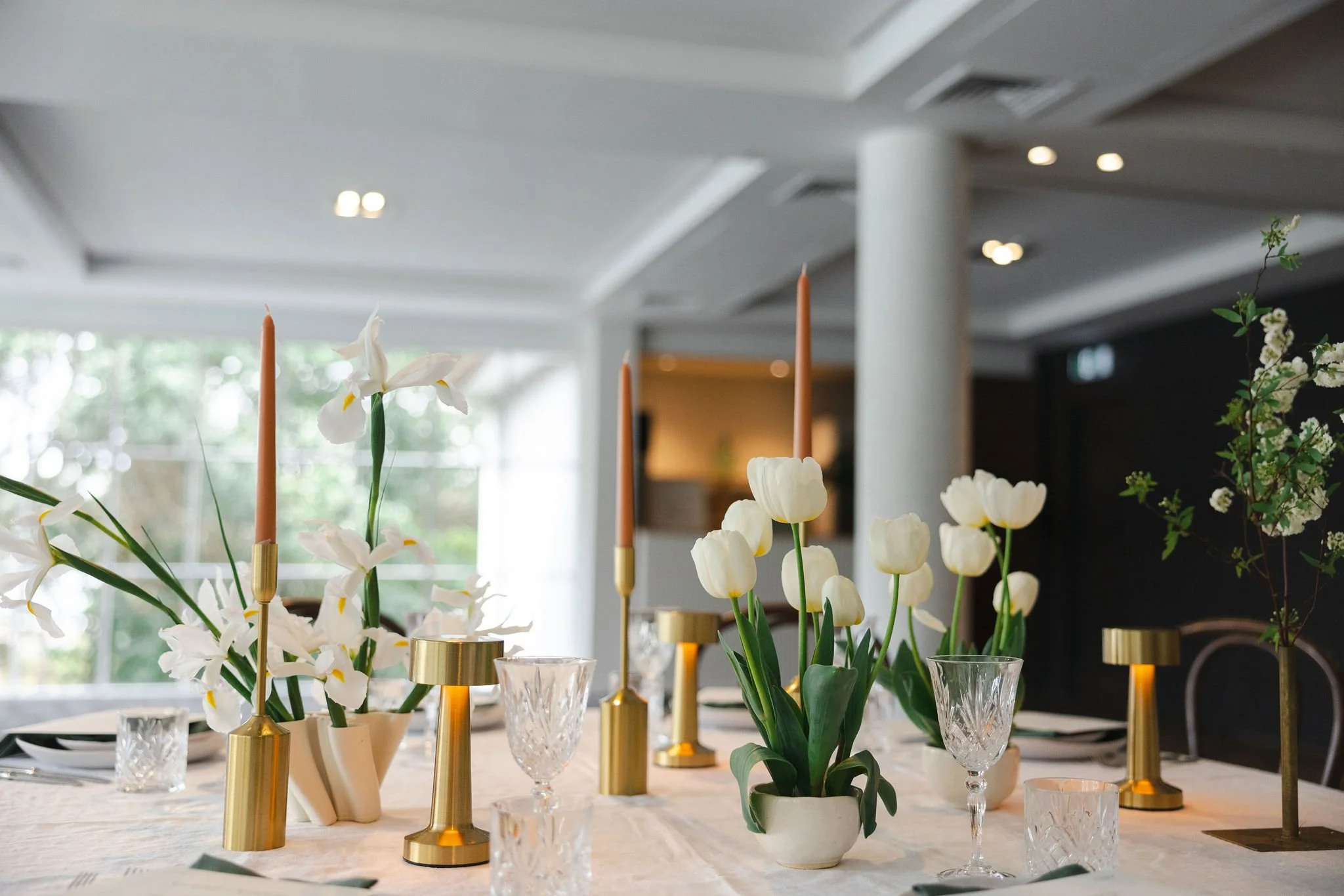 Elegant dining table decorated with white tulips and lilies, gold candleholders with pink taper candles, crystal wine glasses, and tableware in a modern, well-lit room.