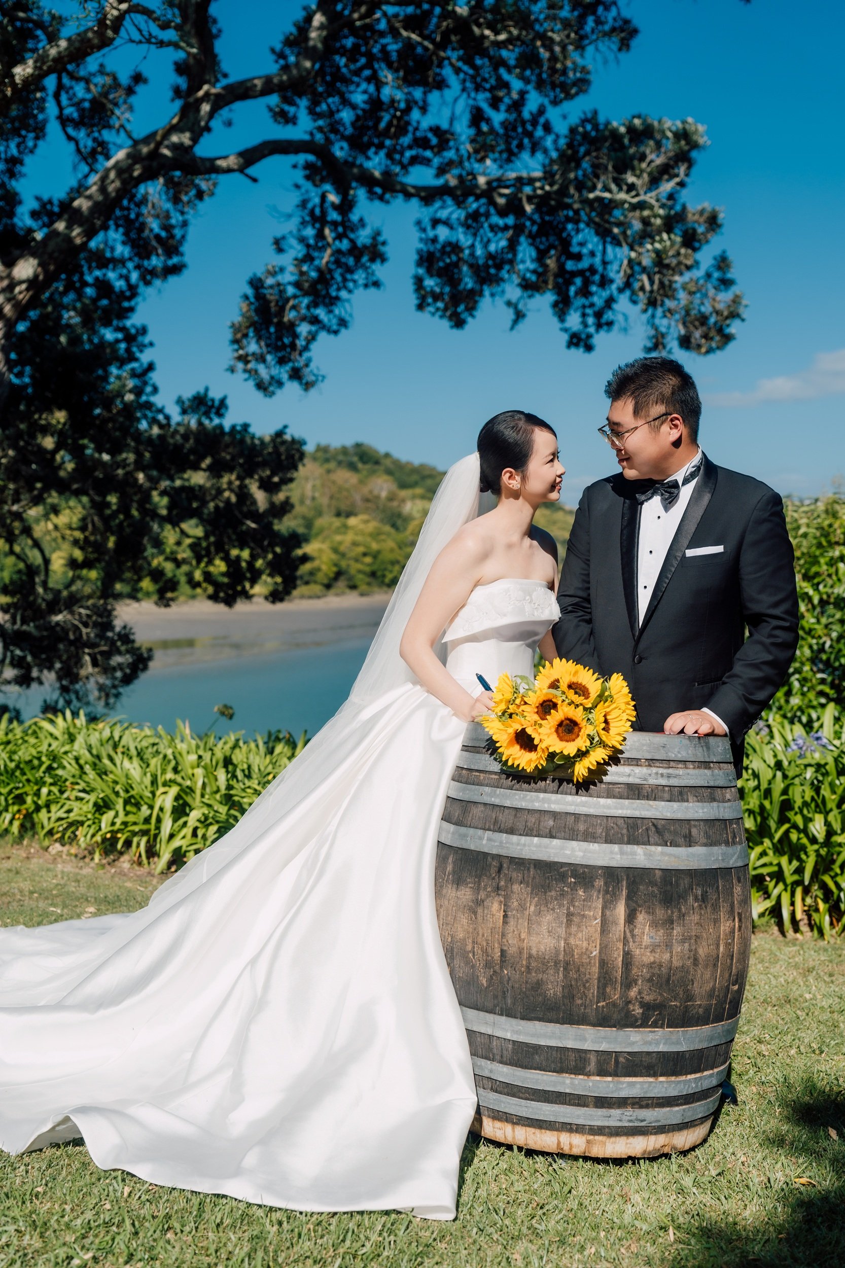 A bride and groom in formal wedding attire standing outdoors next to a barrel with sunflowers, near the water with trees in the background under a blue sky.