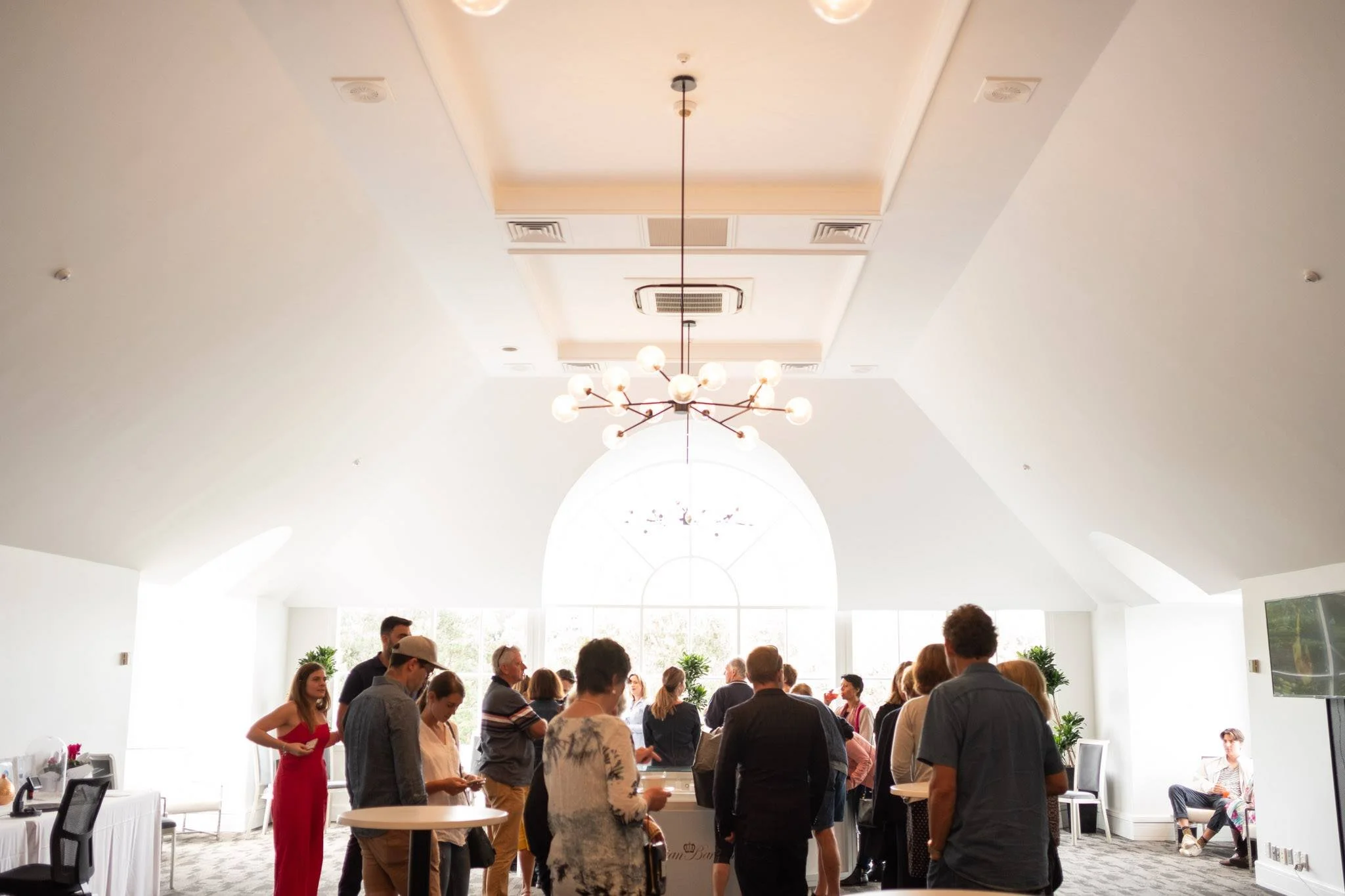 People gathered in a bright, spacious room with a high ceiling, large arched window, and modern chandelier.