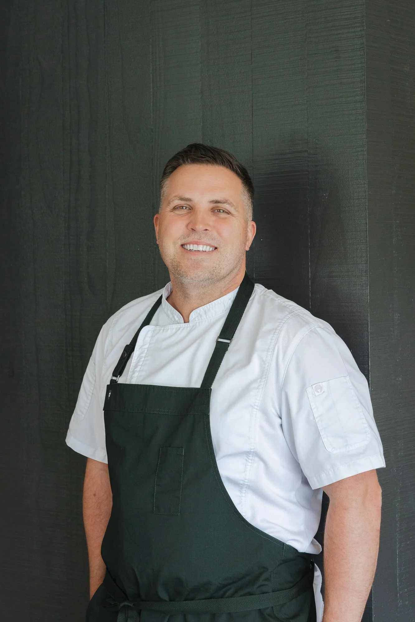 A smiling chef wearing a white chef's jacket and black apron standing against a dark textured wall.