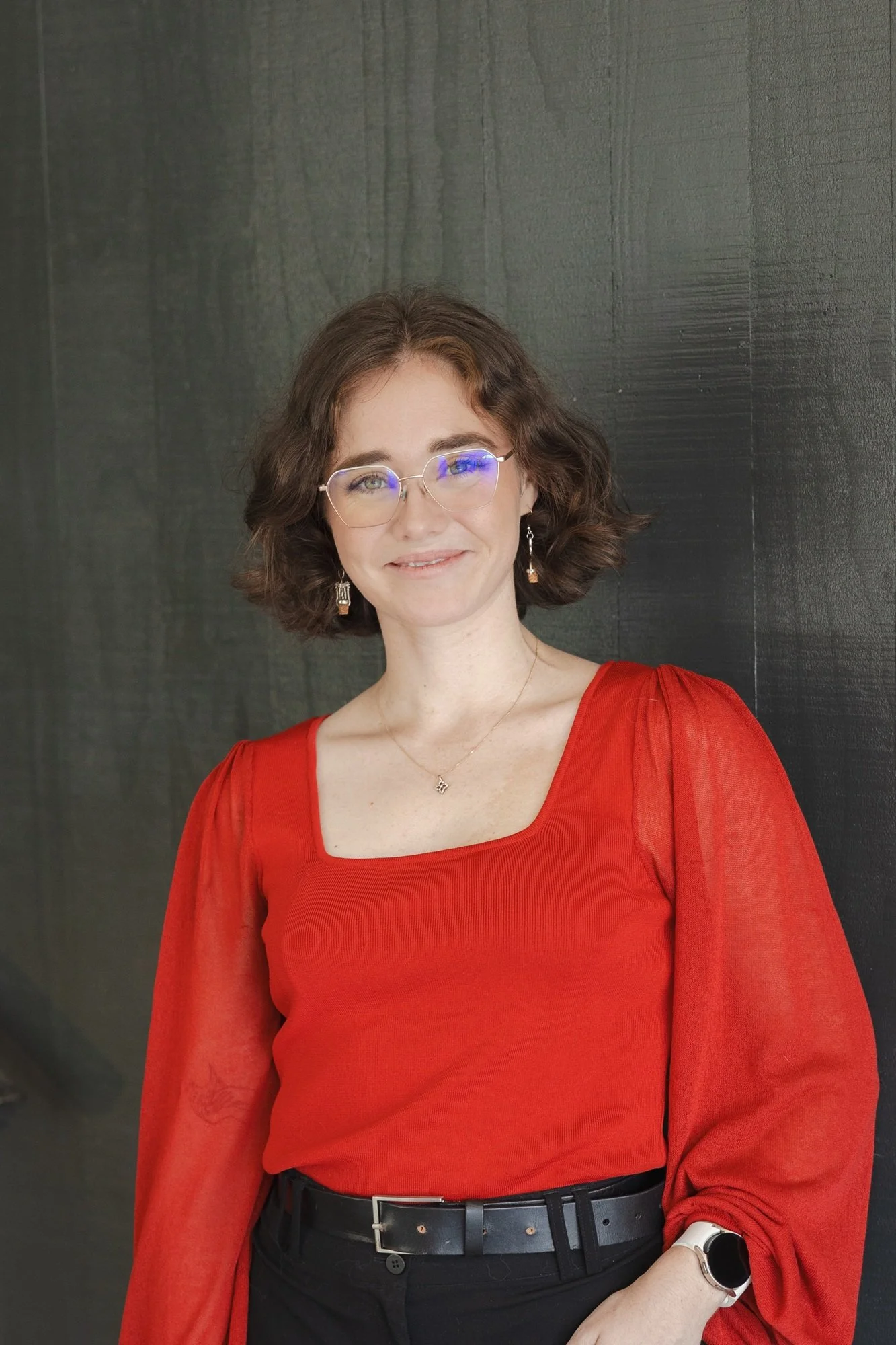 Woman with curly brown hair, glasses, and light skin, wearing a red blouse with sheer sleeves, black pants, a black belt, a necklace, earrings, and a smartwatch, standing against a dark textured background.