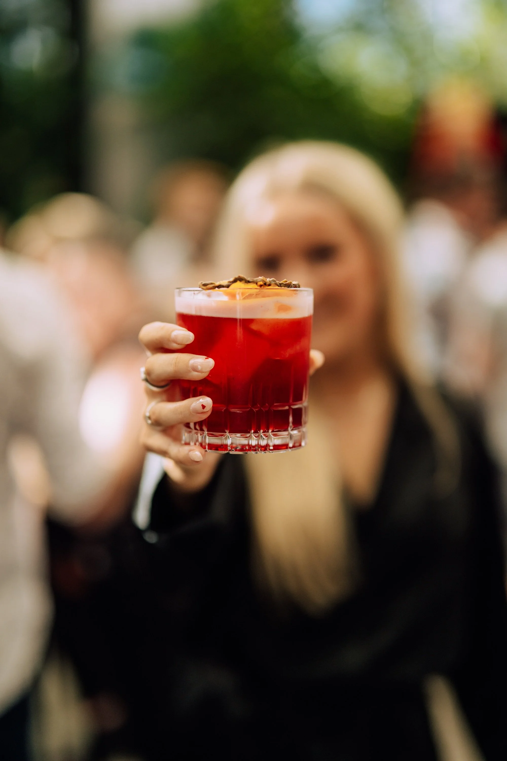 A woman with long blond hair holding up a glass of red cocktail with an orange slice garnish, in focus, with blurred people in the background.