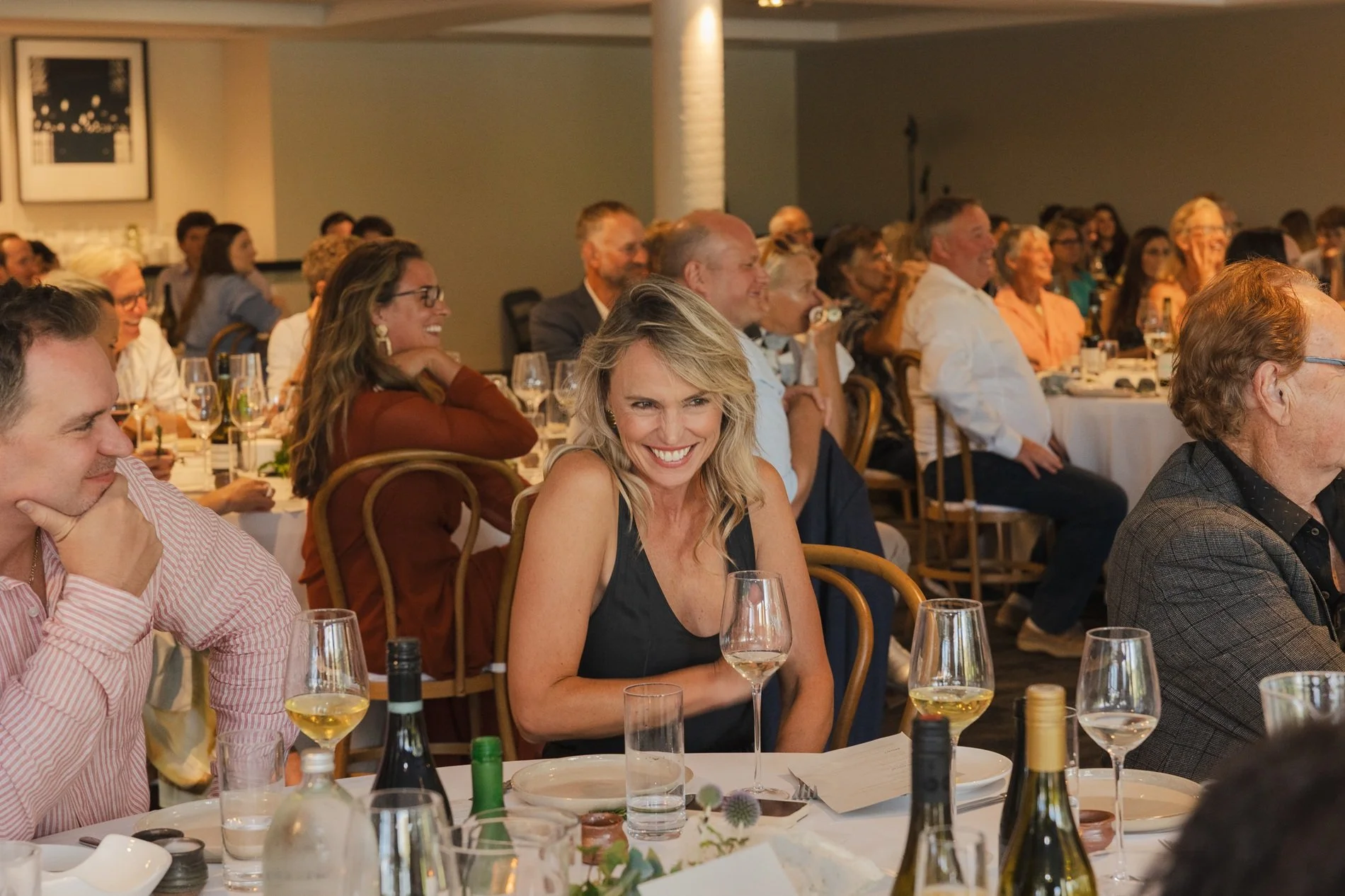 A woman with blonde hair wearing a black dress is smiling at a formal event or dinner, surrounded by other attendees seated at tables with wine glasses and bottles.