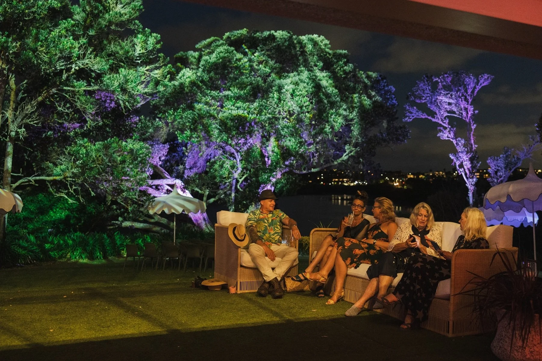 People sitting on an outdoor sofa and chairs on a patio at night, with large trees illuminated in colorful lights in the background.