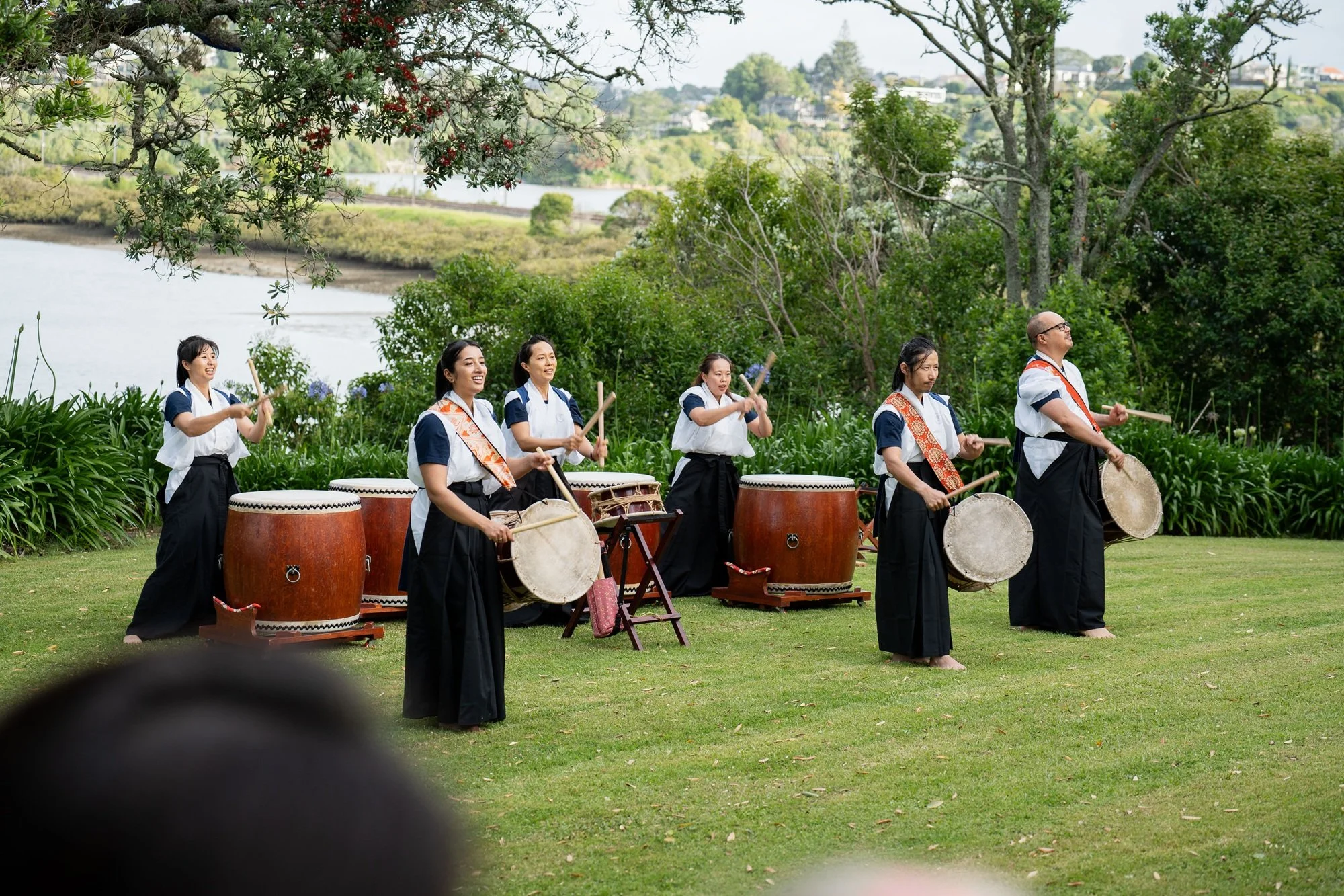 A group of six musicians playing large drums outdoors in a grassy area with trees and a body of water in the background.