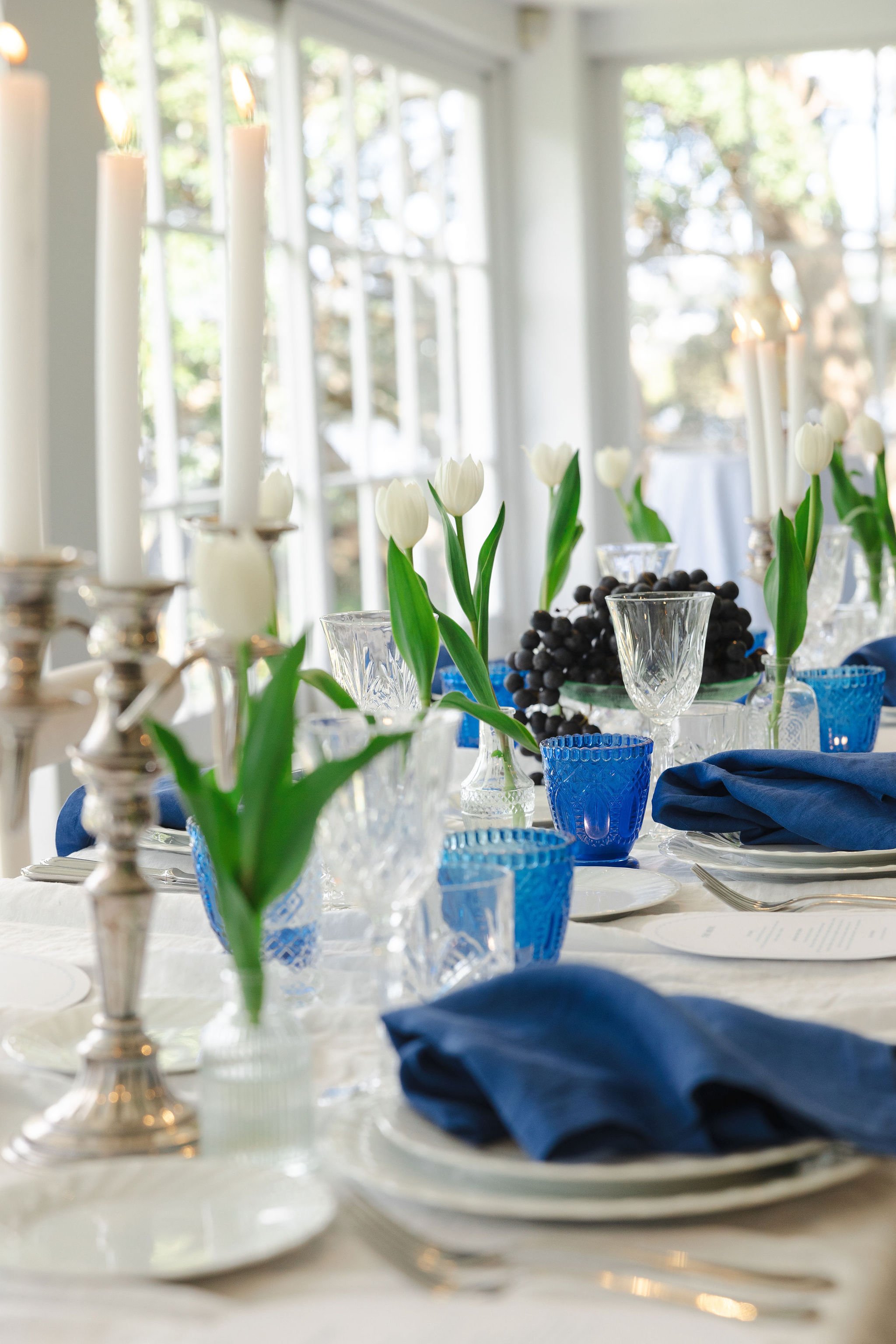 Decorated dining table with white tulips in vases, candlesticks, blue glassware, and a centerpiece of black grapes.