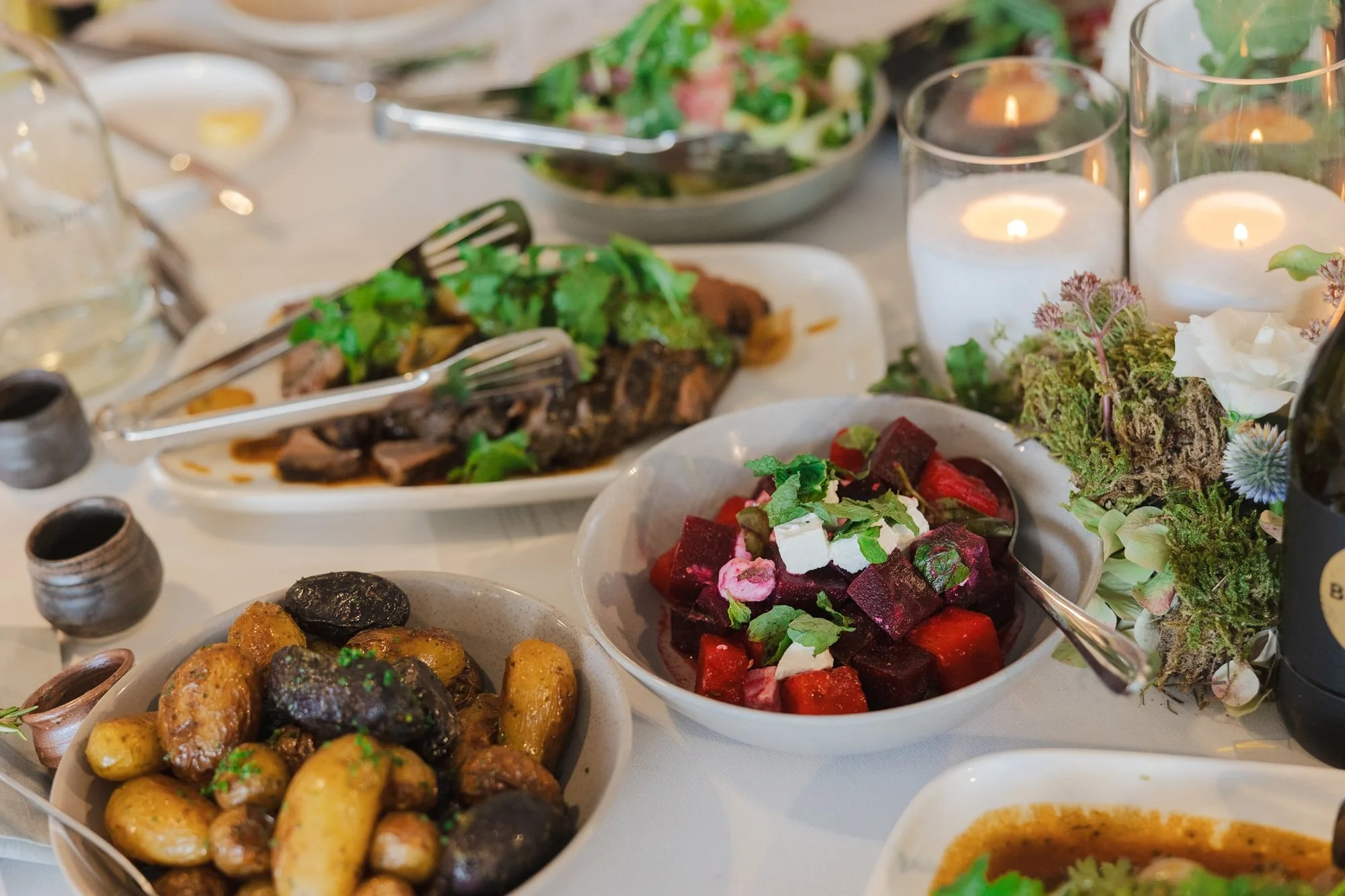 A table set with various dishes including roasted vegetables, beet salad with feta, a plate of cooked meat with greens, and candles as decoration.