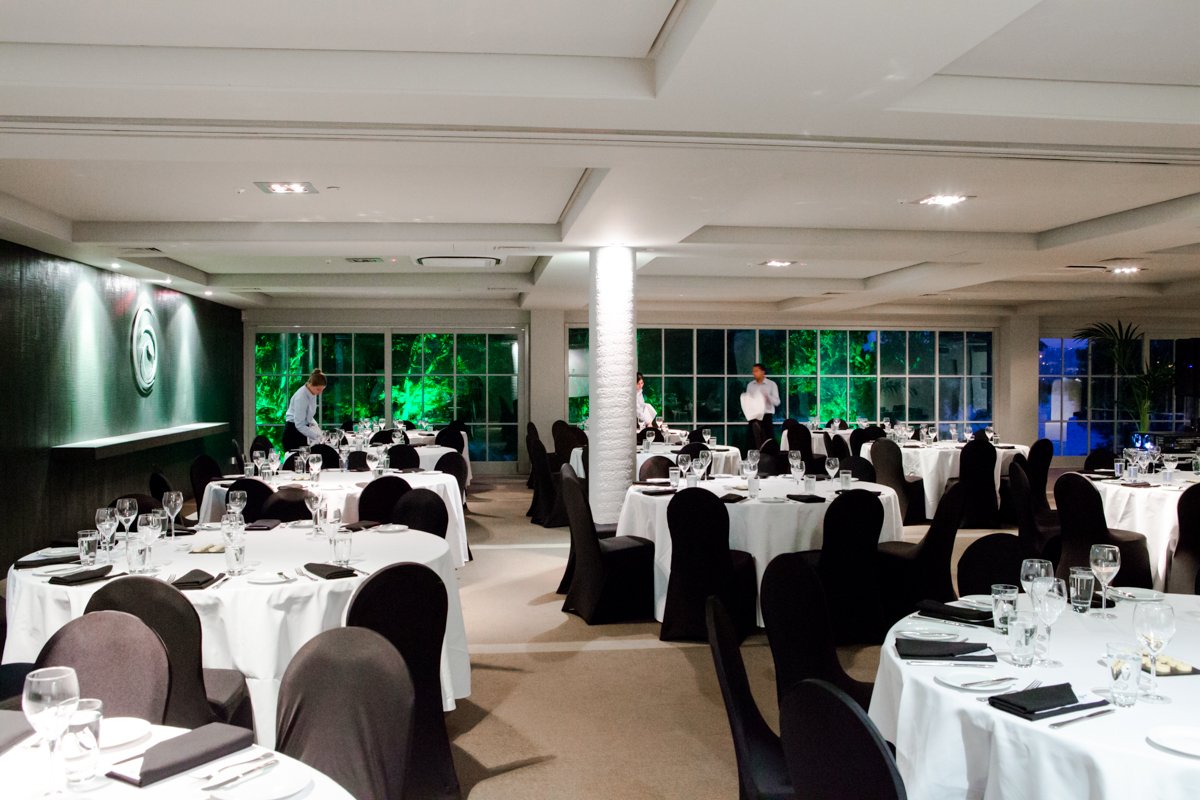 Empty banquet room with round tables covered in white tablecloths, set with glassware, silverware, and folded black napkins, with waitstaff preparing the space and large windows showing greenery outside.