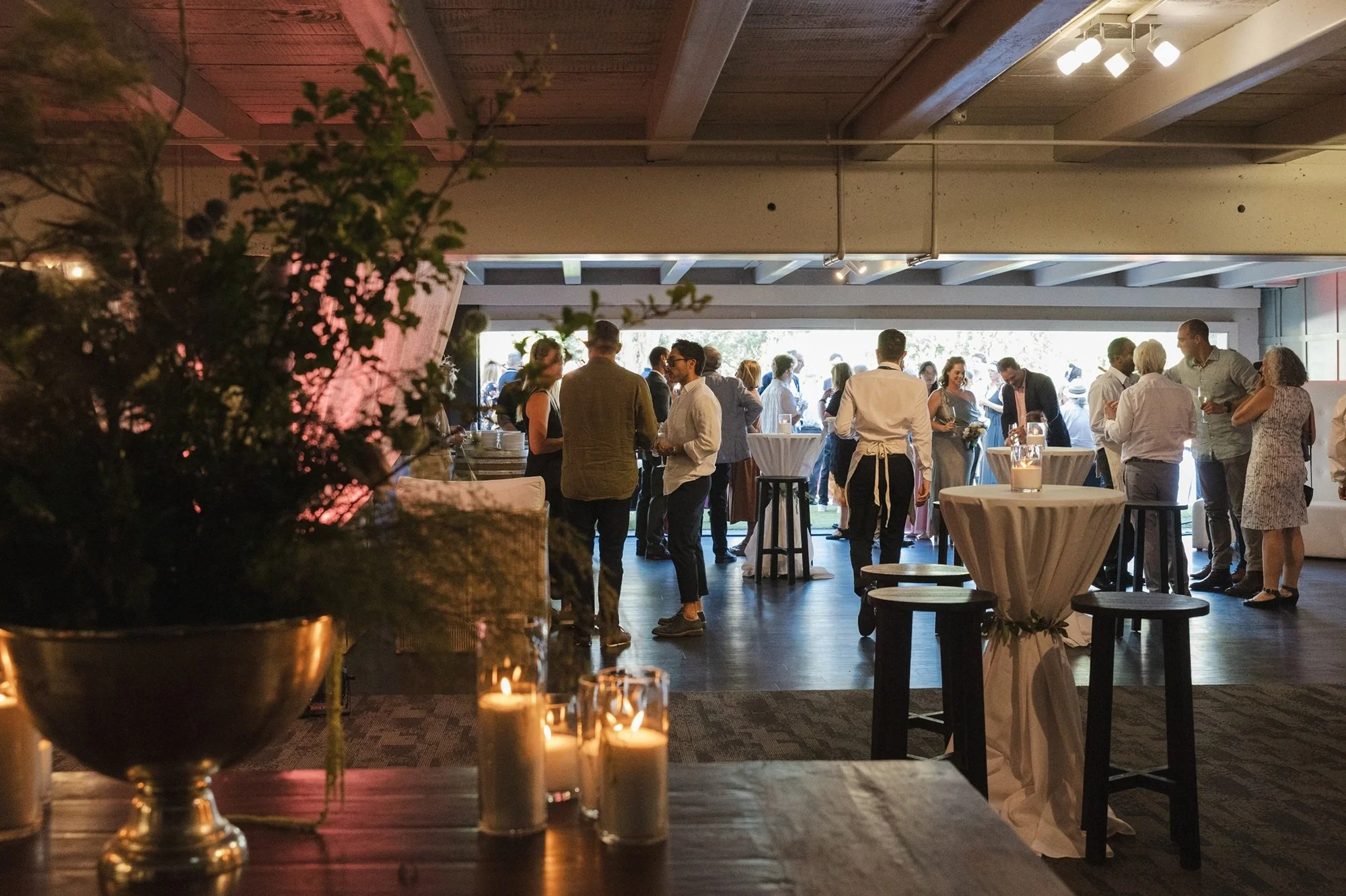 Guests socializing at a reception with high tables and candles, under a modern indoor venue with large open entrance and natural light.