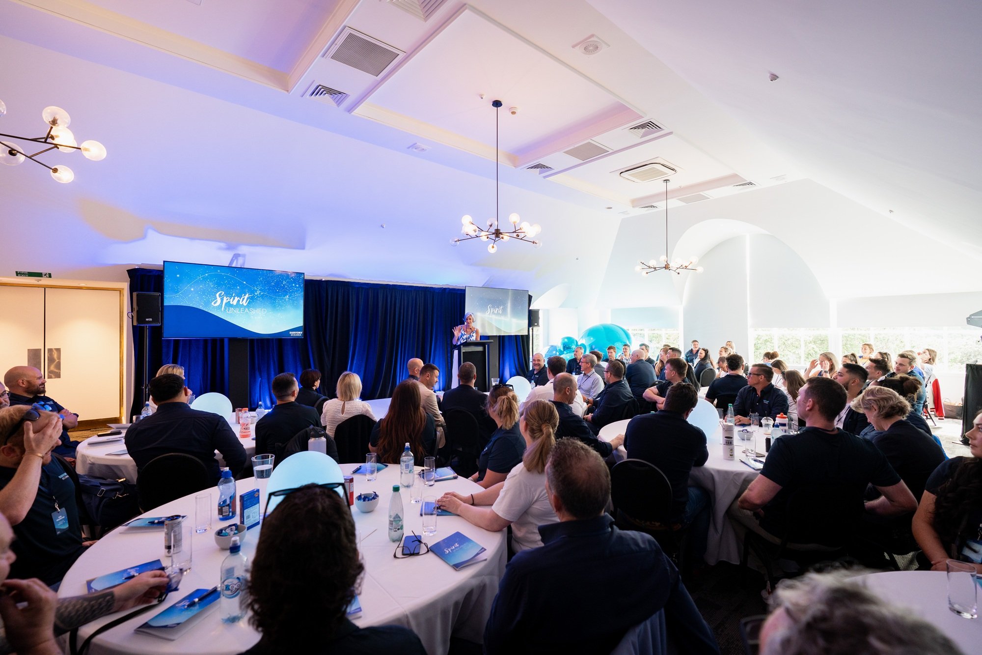 Conference room filled with people seated at round tables, listening to a speaker on stage with screens displaying 'Spirit Unleashed' in the background, decorated with blue balloons and balloons on tables, bright natural light coming through large windows.