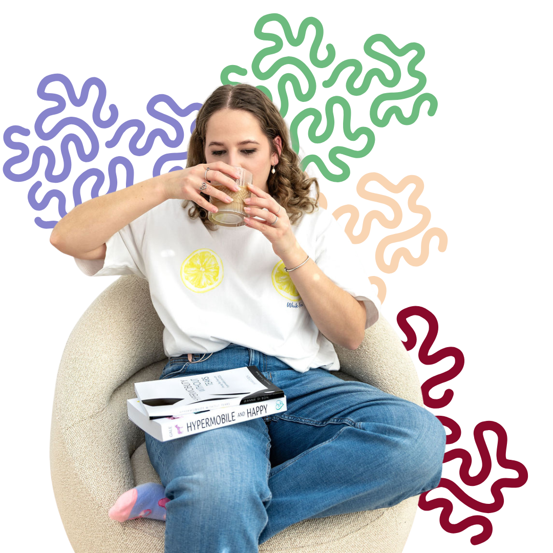 Young woman with curly hair sitting on a beige armchair, drinking from a glass. She is wearing a white T-shirt with lemon prints, blue jeans, and pink socks. Books are resting on her lap.