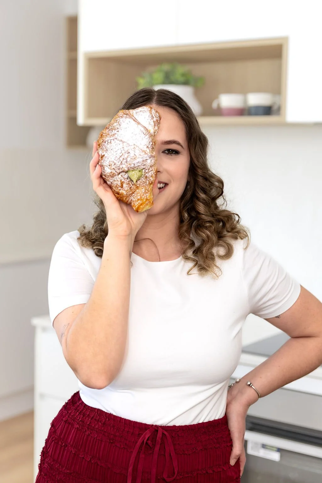 A woman with curly brown hair smiling and holding a large piece of pastry over her right eye in a modern kitchen.