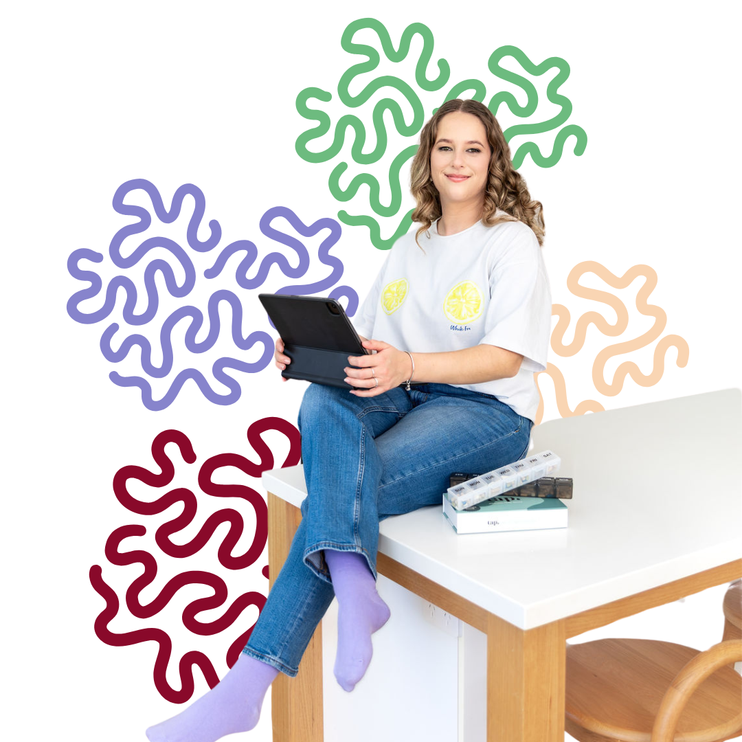 A young woman with wavy light brown hair and fair skin, sitting on a white table with wooden legs, wearing a white t-shirt with lemon graphic, blue jeans, and purple socks, holding a tablet. She is smiling and looking at the camera. Next to her on the table are a transparent pill organizer, a black case, and a closed book. The background features abstract colorful shapes in purple, green, orange, beige, and maroon.