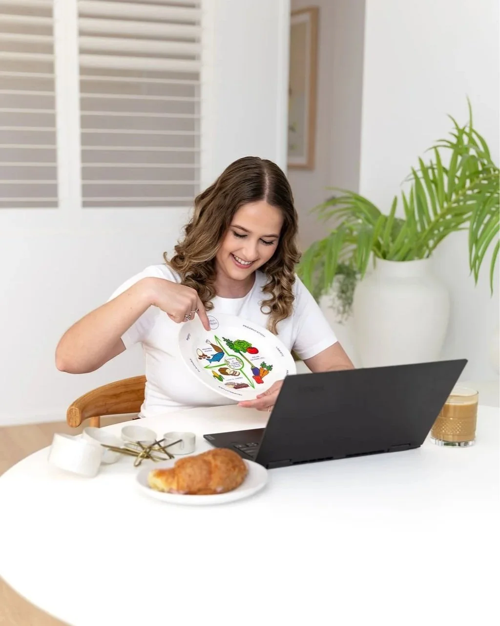 Woman sitting at a table, smiling and pointing at a diagram on a plate, with a laptop, food, and drink on the table, in a bright, modern room with plants.