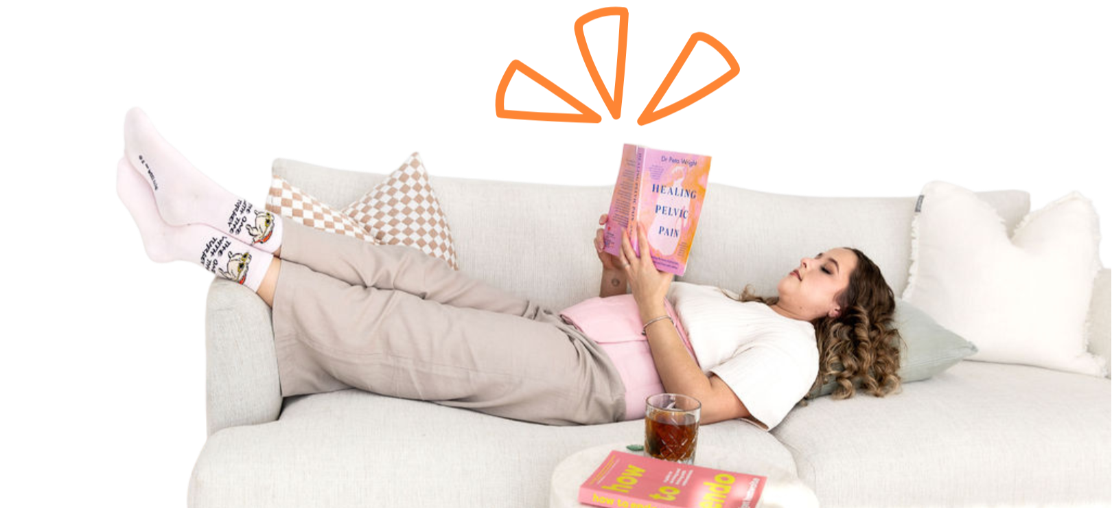 Woman lying on a beige sofa reading a book titled "Healing Pelvic Pain" with a glass of dark beverage and a pink book on the table beside her, wearing socks with cartoon characters.