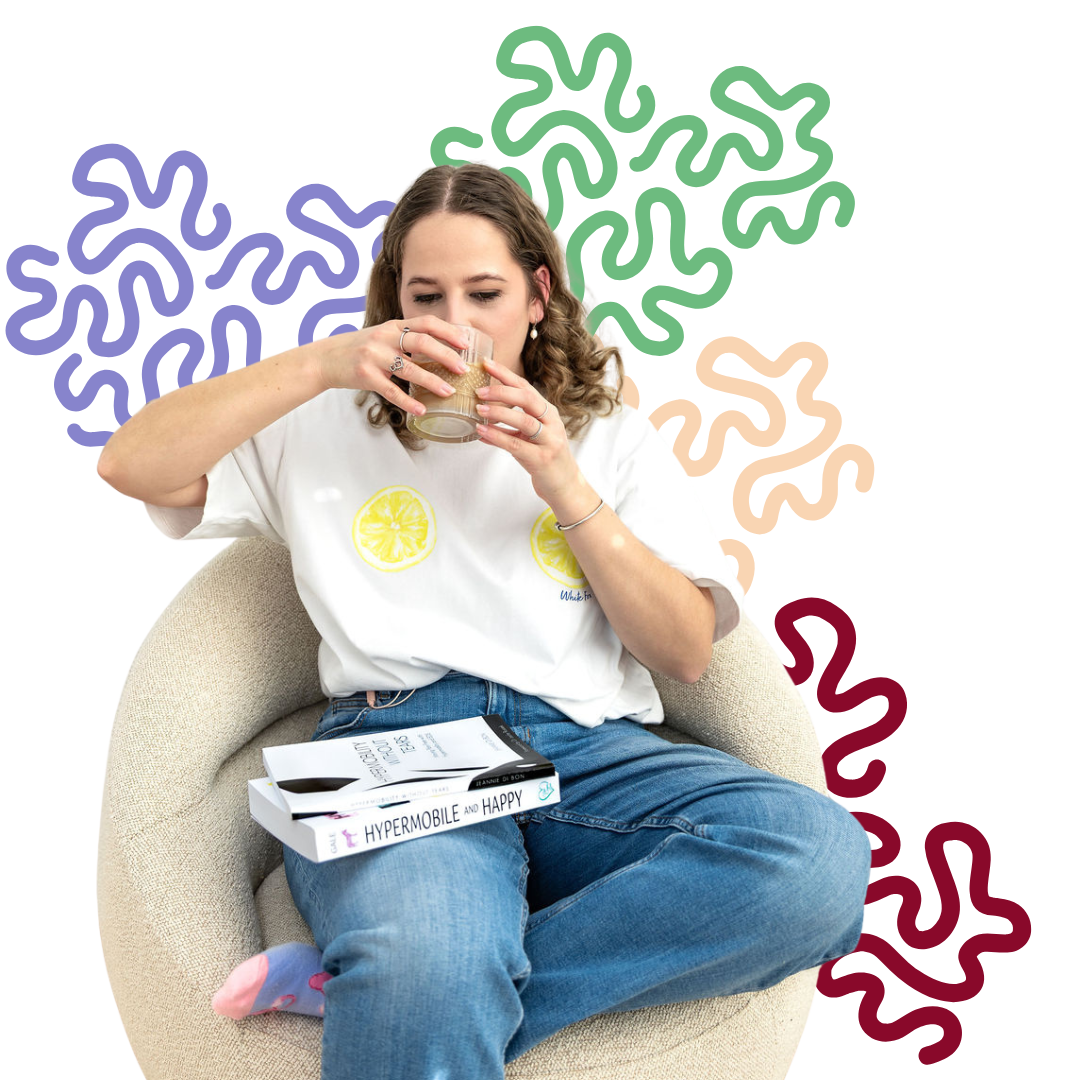 A woman sitting in a beige armchair drinking a beverage from a glass, with books on her lap, wearing a white t-shirt with lemon slices, blue jeans, and pink socks.