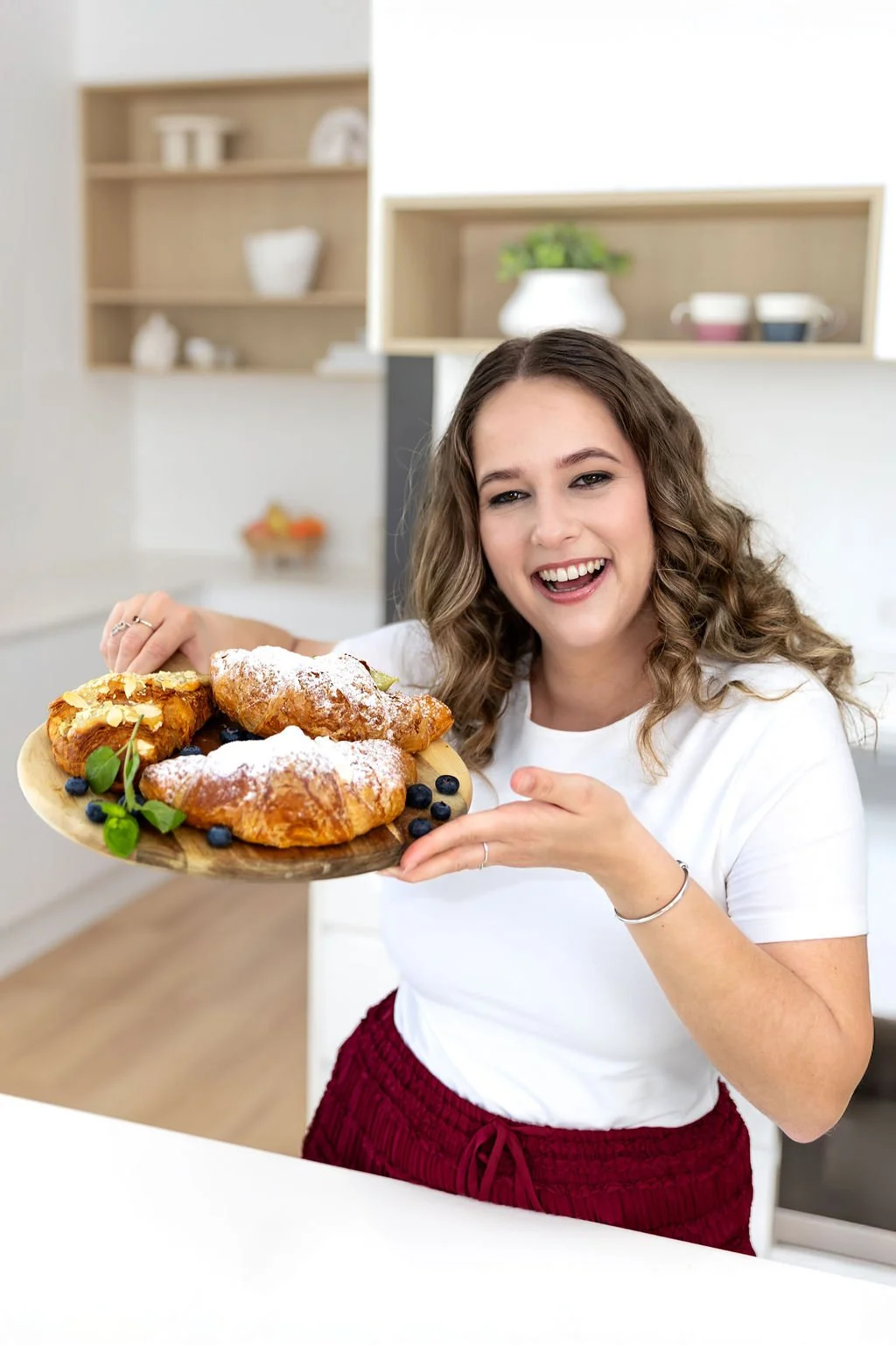 A woman with curly hair holding a wooden plate with pastries topped with powdered sugar and garnished with blueberries and mint leaves in a modern kitchen.