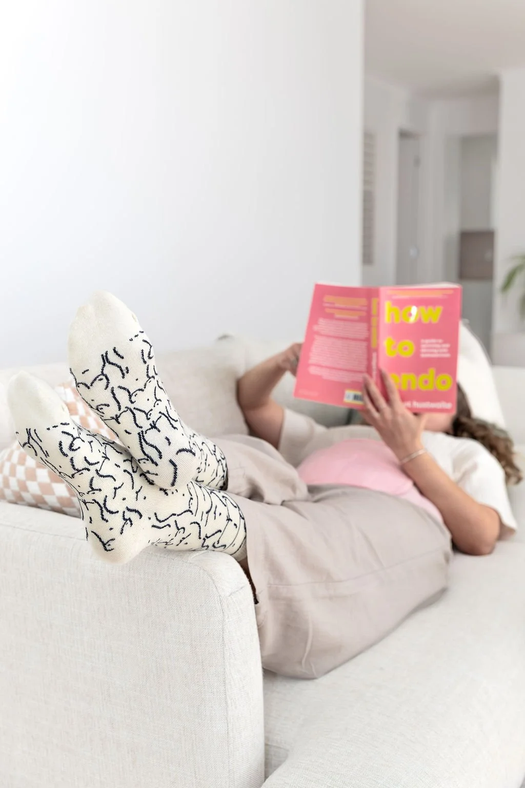 Person lying on a cream-colored sofa with feet up, wearing white socks with black squiggly lines, and reading a pink book titled 'How to' in a bright, modern living room.