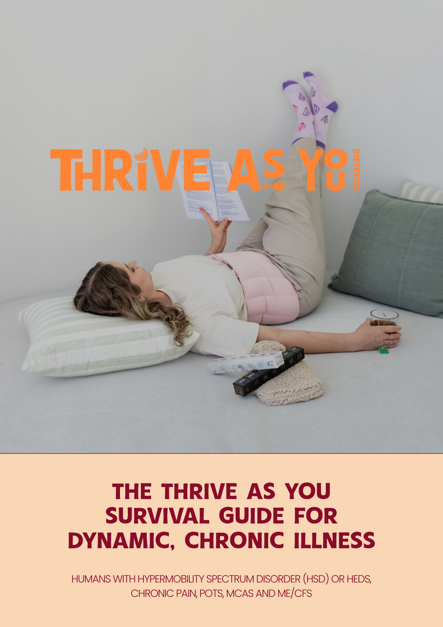 A woman lying on her back on a bed, reading a brochure with a glass of beverage in her hand. She is wearing socks with purple accents, beige pants, and a white shirt. The bed has a striped pillow and a green pillow. There are extension cords and a cozy blanket beside her. The image features the text "Thrive As You" in large orange letters and a subtitle "The Thrive As You Survival Guide for Dynamic, Chronic Illness" in bold maroon text, with additional smaller text about humans with hypermobility spectrum disorder, chronic pain, and related conditions.