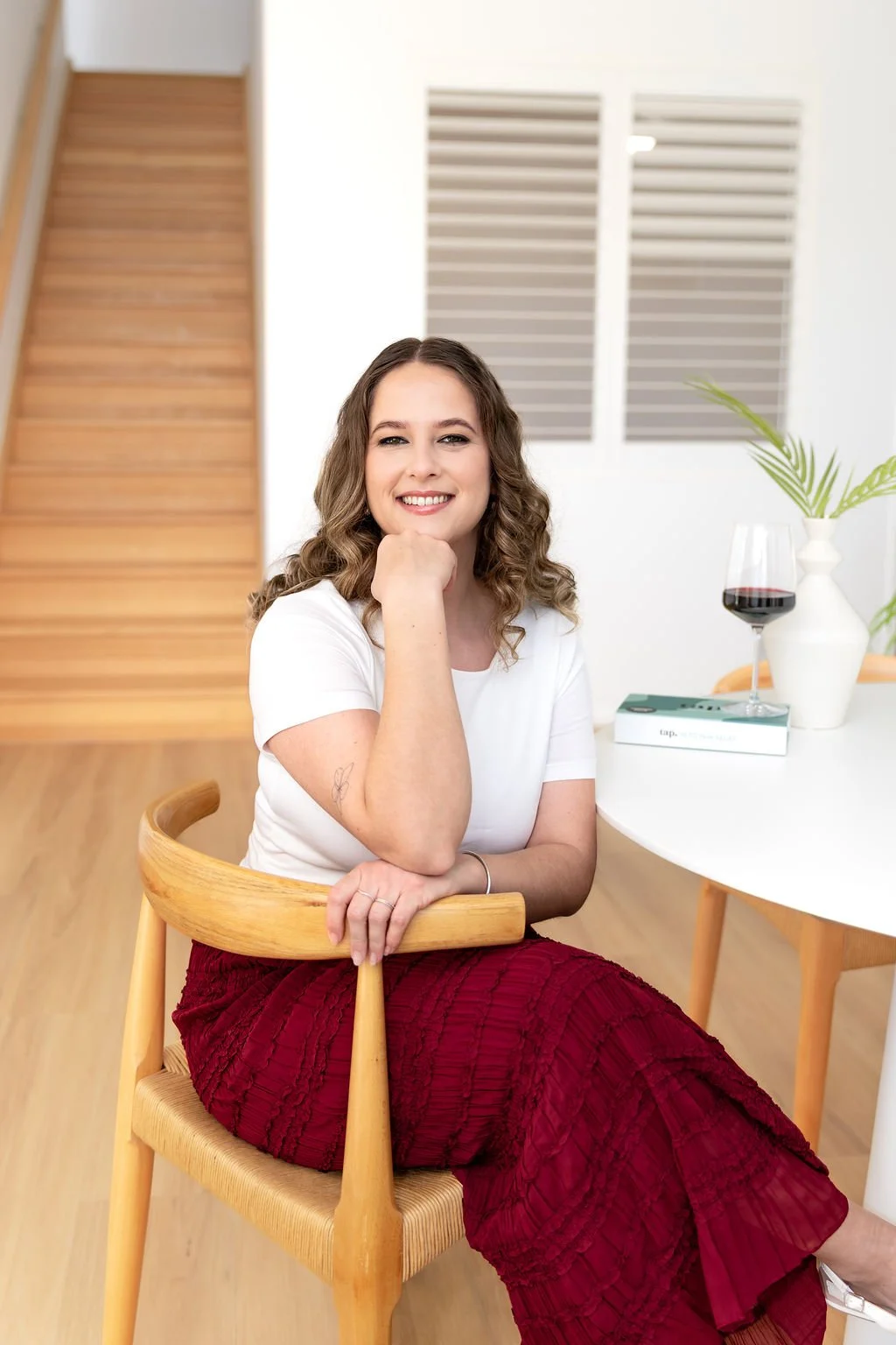 A woman with curly brown hair and a bright smile sitting at a white table with a glass of red wine, a white vase with green leaves, and a closed book. She is wearing a white top and a red skirt, and has a tattoo on her left arm. The background features a wooden staircase and white wall with windows.