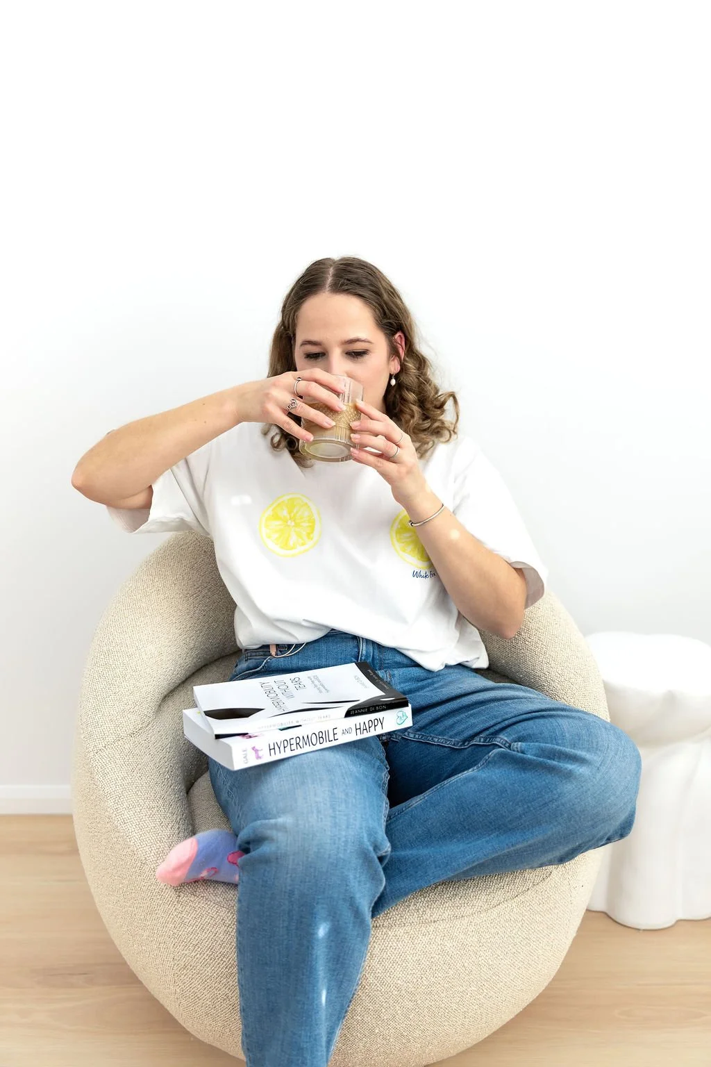 Woman sitting in a beige chair drinking from a glass, with a book titled 'HYPERMOBILE AND HAPPY' resting on her lap, wearing a white T-shirt with lemon slices print and blue jeans, in a minimal white room.