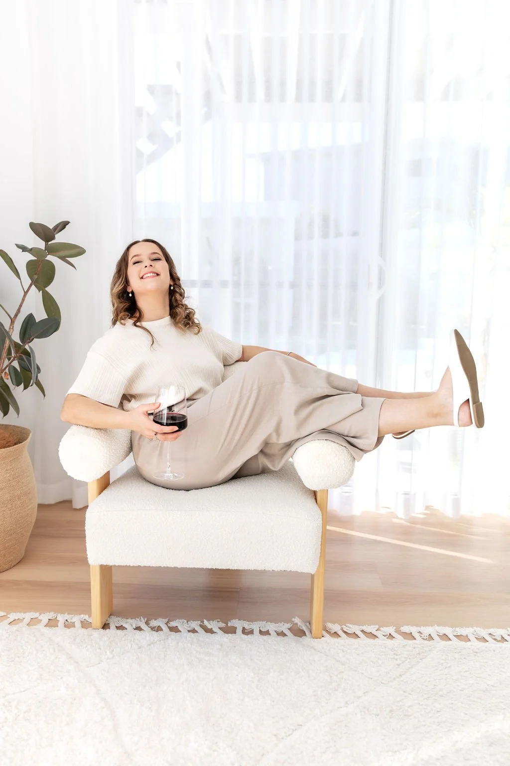 A woman sitting comfortably on a small sofa, holding a glass of red wine, smiling with her legs raised and crossed at the ankles, in a bright room with sheer white curtains and a potted plant nearby.