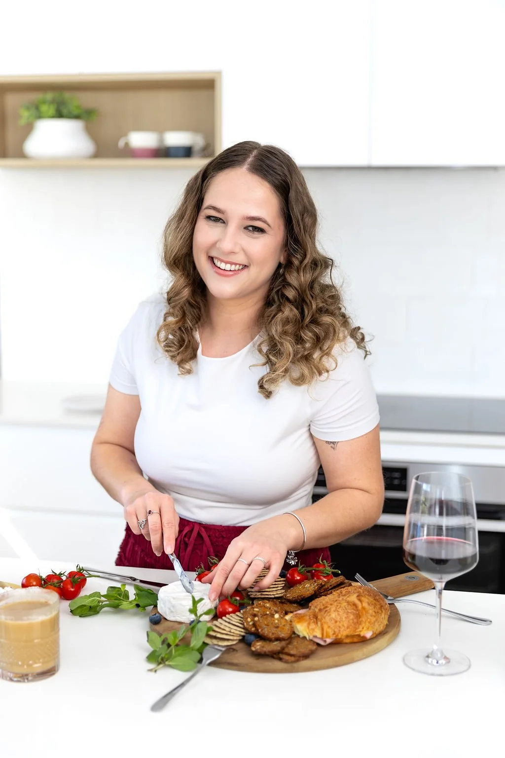 A woman with curly brown hair and a white shirt, smiling and cutting cheese, standing at a kitchen counter with a cheese platter, cherry tomatoes, and a glass of red wine.