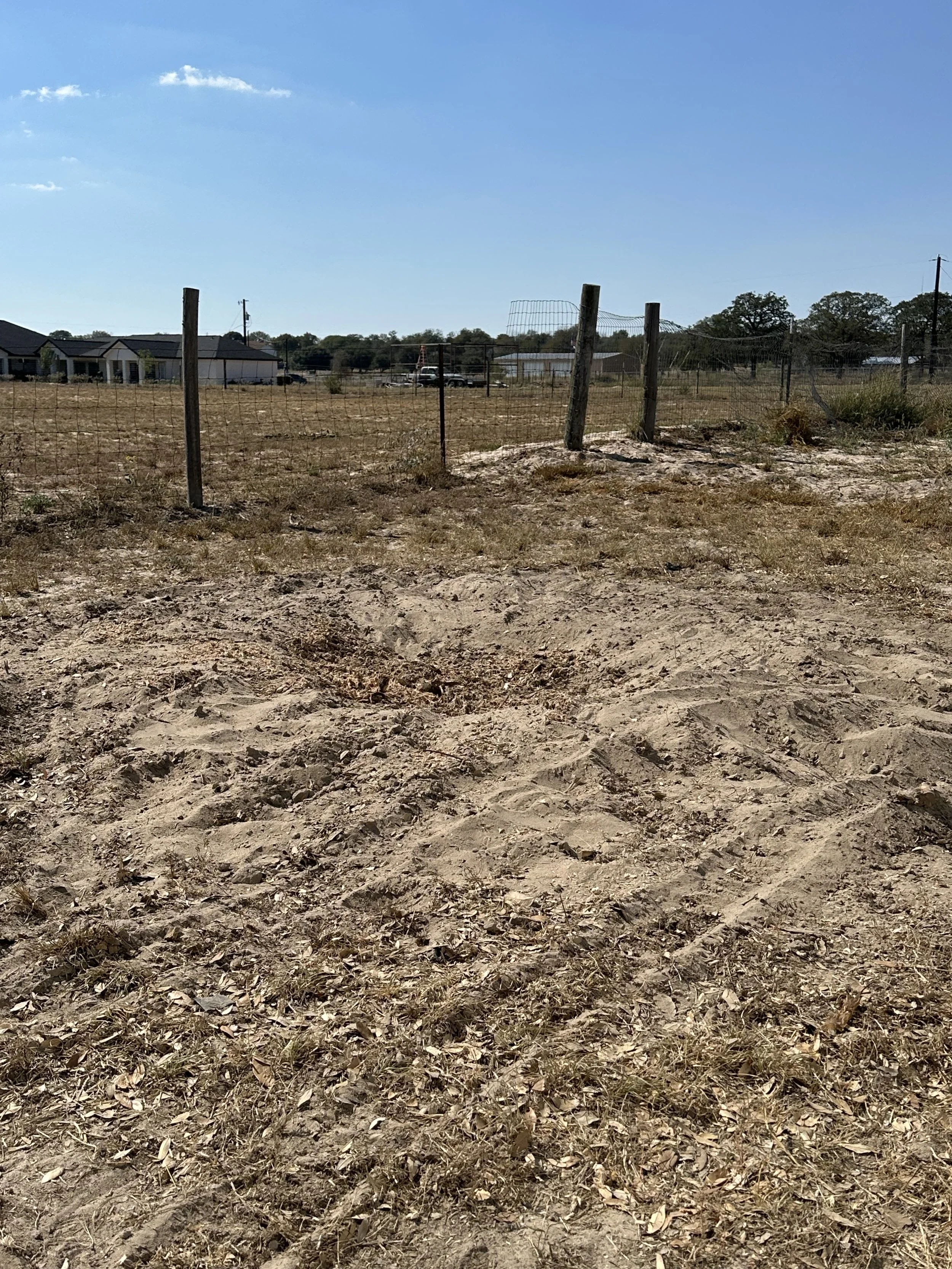Dirt area with a wire fence and wooden posts in a rural setting, homes and trees in the background, under a bright blue sky with a few clouds.