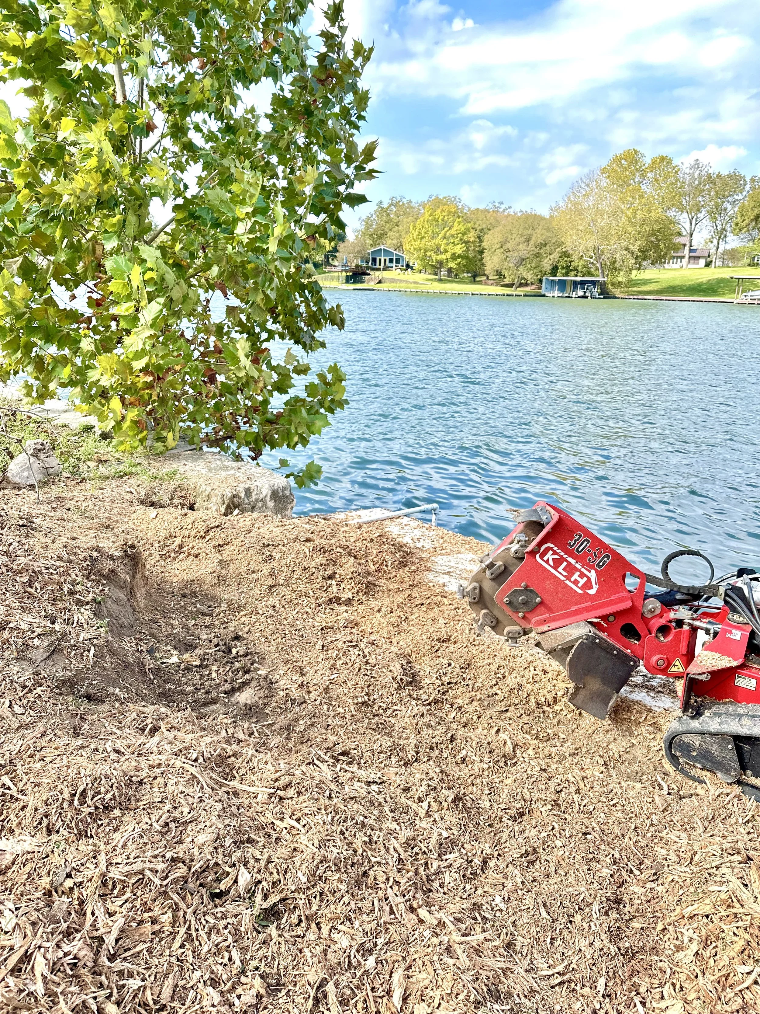 A lakeside scene with a red landscaping power tool on the ground, a large leafy tree, and a calm body of water. In the background, there are houses and trees on the opposite shore under a partly cloudy sky.