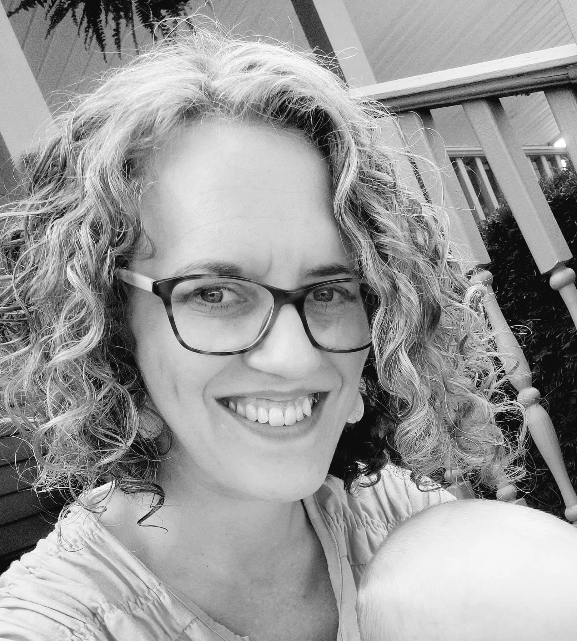 Close-up of a woman with curly hair, glasses, and a smile, taken outdoors on a balcony with a railing and plants in the background, in black and white.