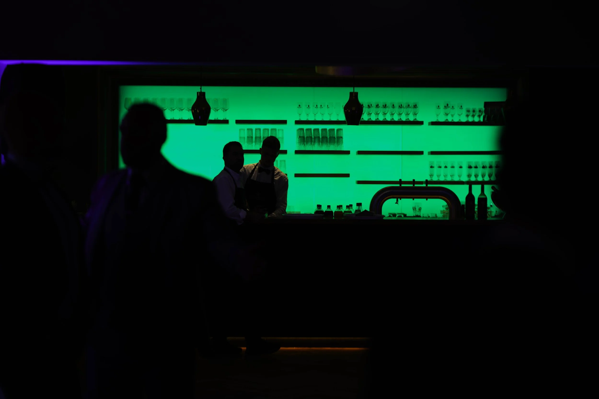 Dunkler Blick in eine Bar mit grüner Beleuchtung, silhouettierte Menschen und Barkeeper im Hintergrund.