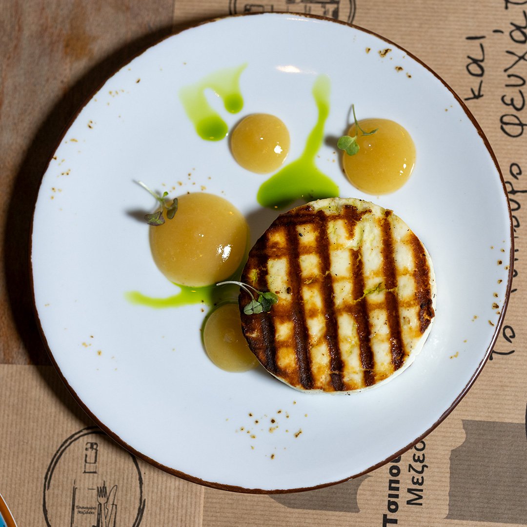 Grilled cheese sandwich with tomato slices, garnished with microgreens and drizzled with green sauce, served on a white plate.
