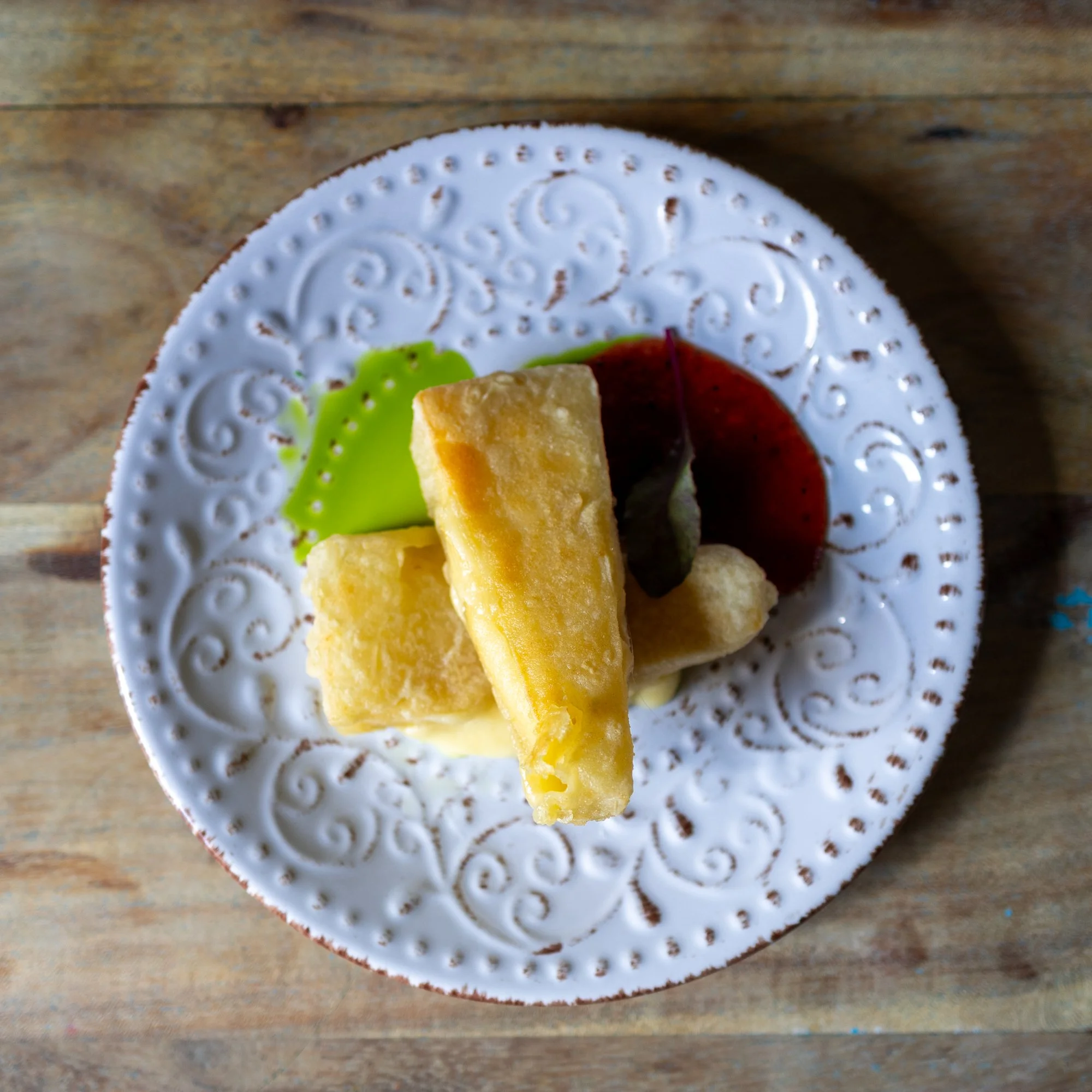 Fried fish fillets served with green sauce, red dipping sauce, and a leaf on a white, embossed plate on a wooden surface.