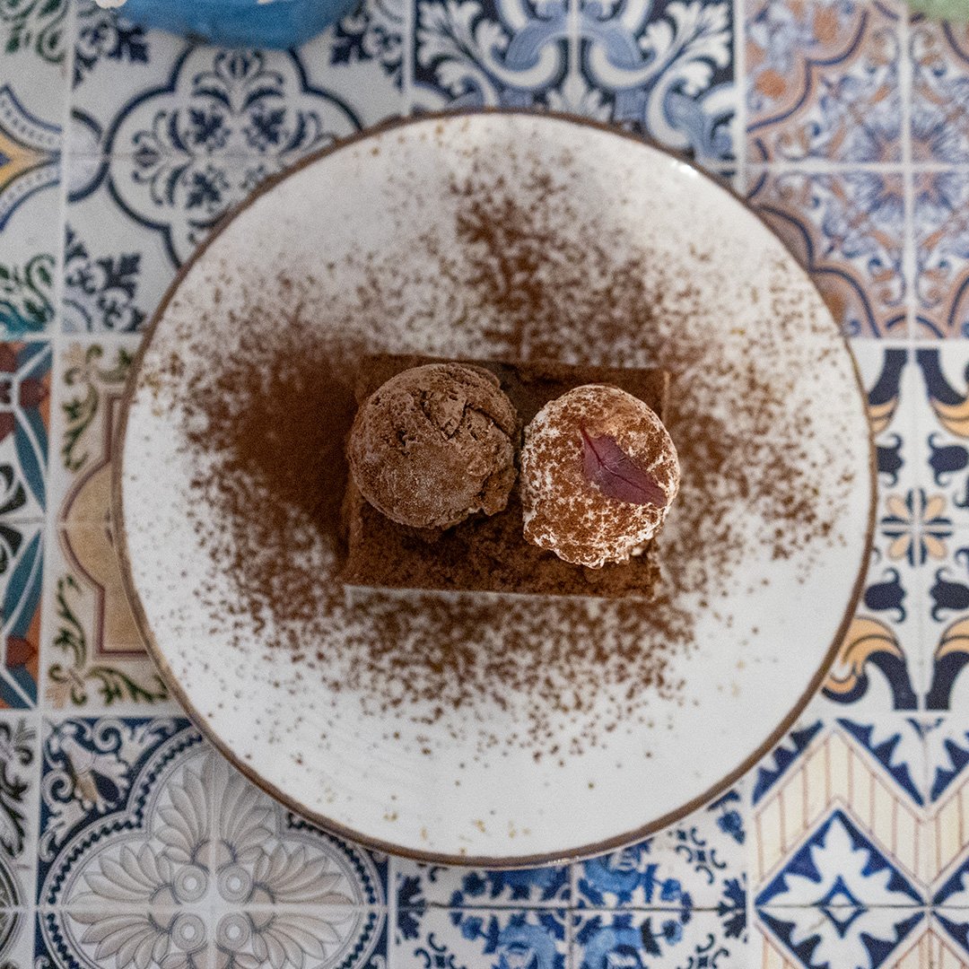 A dessert on a white plate with a decorative brown pattern, featuring a chocolate brownie topped with a dusting of cocoa powder, a chocolate truffle, and a halved truffle with a leaf garnish, set on a colorful patterned tile surface.