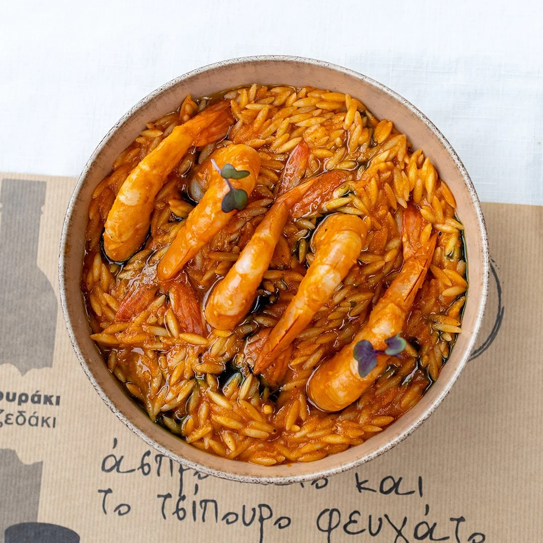 A bowl of seafood orzo pasta with shrimp, garnished with small herbs, on a piece of paper with Greek writing.