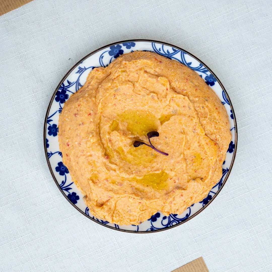 A decorative bowl filled with a creamy, orange-colored dip or spread garnished with a cherry tomato and a small green leaf, placed on a white textured surface.