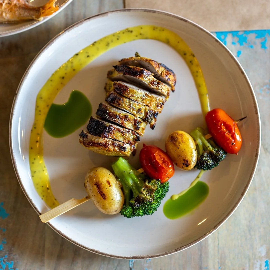 Sliced grilled fish with roasted cherry tomatoes, baby potatoes, broccoli, and sauce garnishes on a white plate.