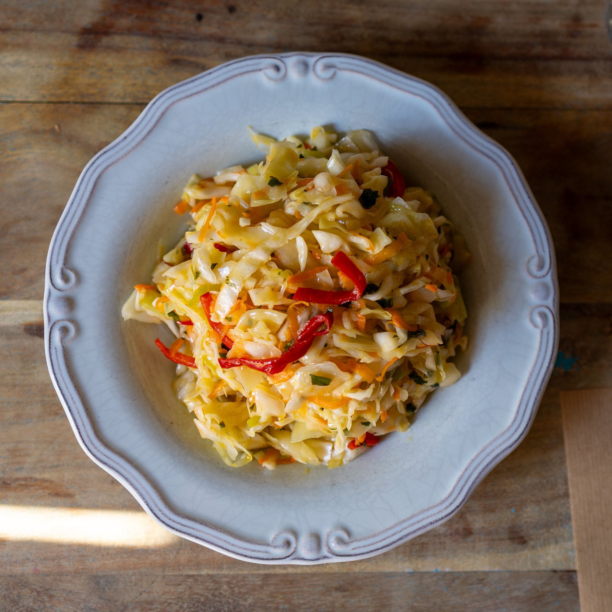 A white decorative bowl containing a serving of stir-fried cabbage with red and orange bell peppers on a wooden surface.