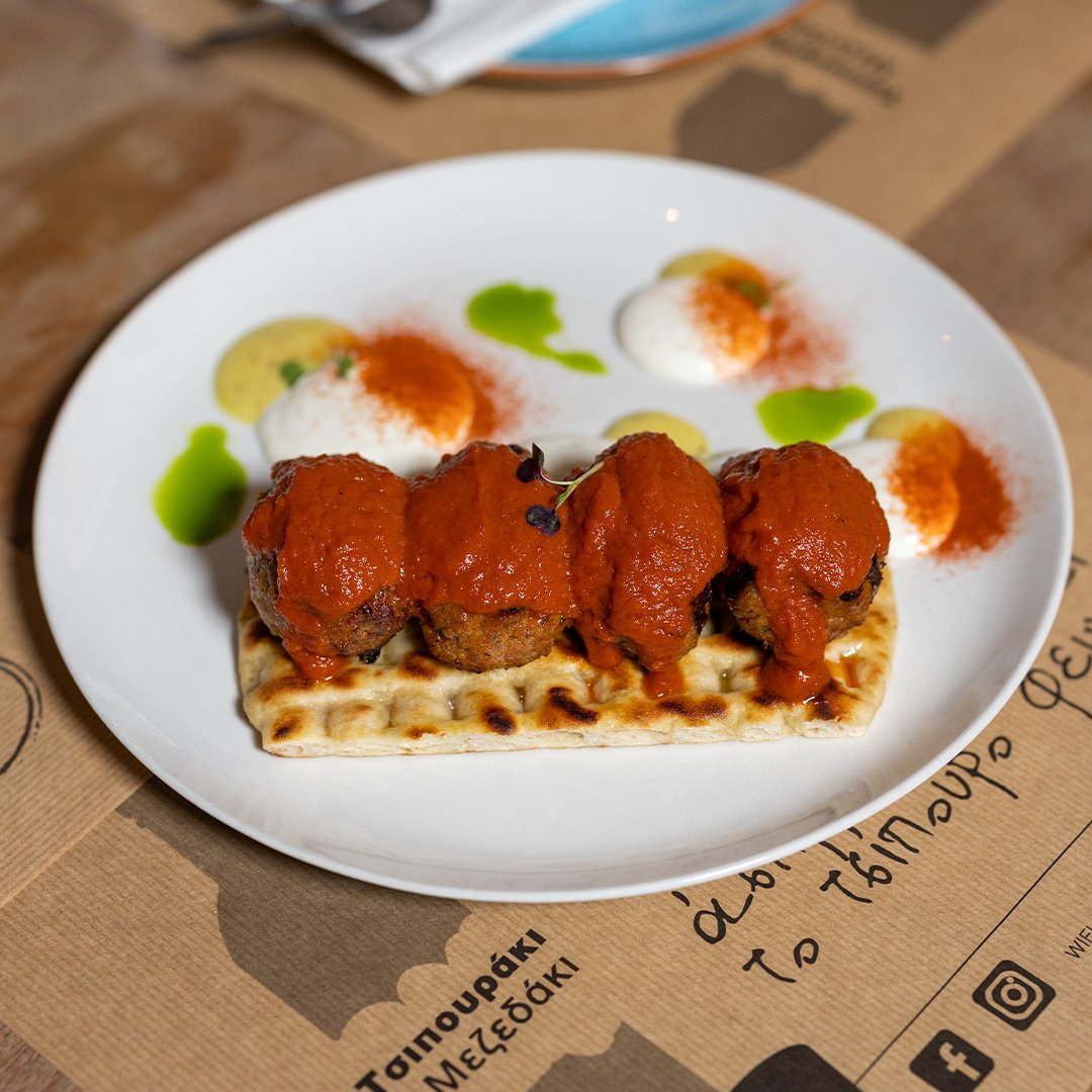 Plate with grilled meatballs topped with tomato sauce, served with a slice of flatbread, and accompanied by dollops of white sauce garnished with red paprika, green sauce, and pepper.