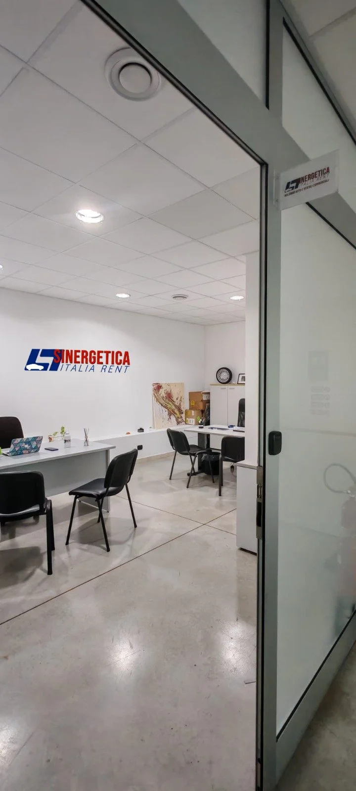 Interno di un ufficio con pareti bianche, scrivanie, sedie nere, un armadietto bianco e un logo sulla parete che dice Sinergética Italia Rent.