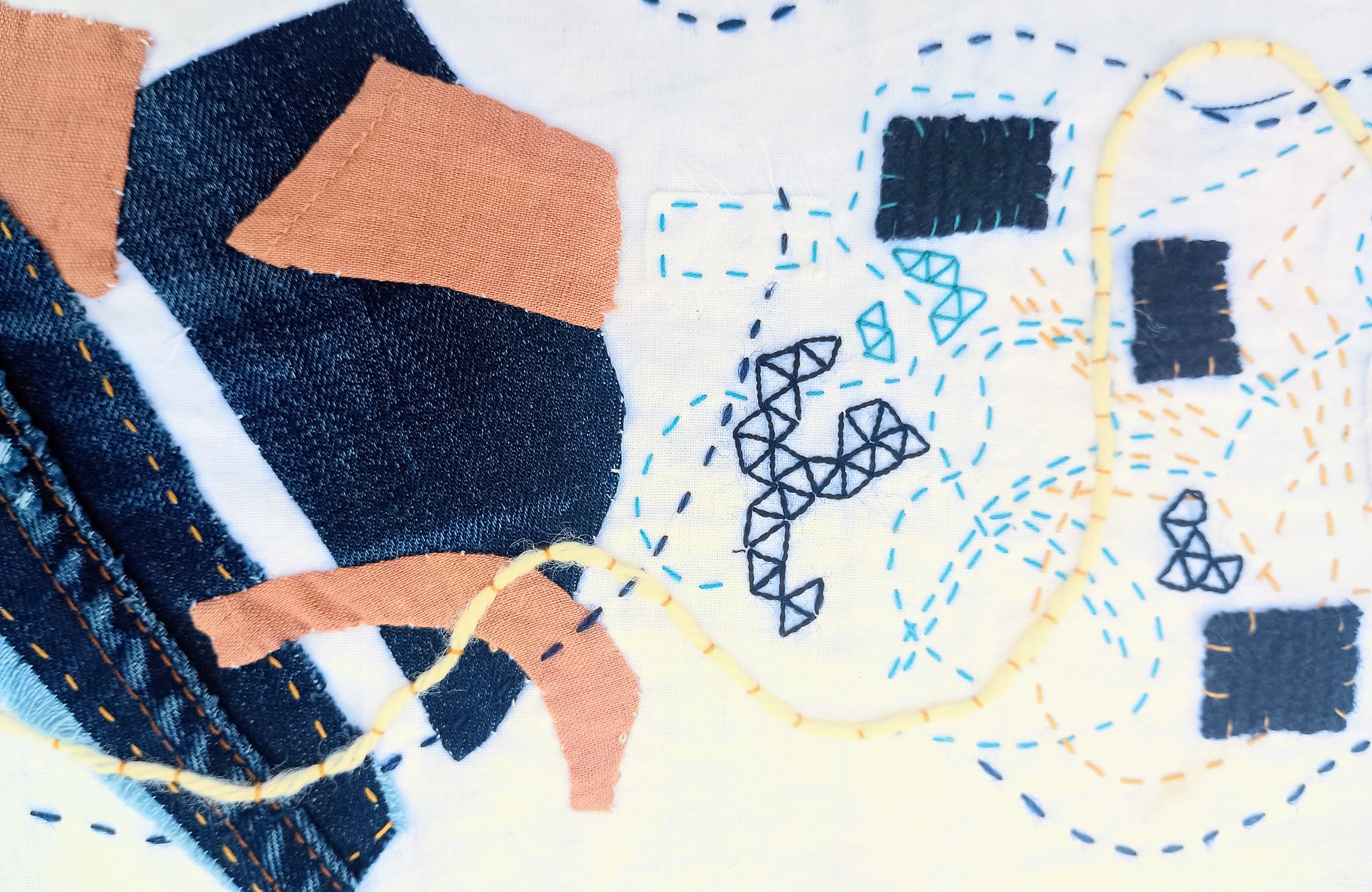 Close-up of fabric with embroidered designs, including denim material with orange and black patches, yellow stitched lines, and black embroidered geometric shapes.