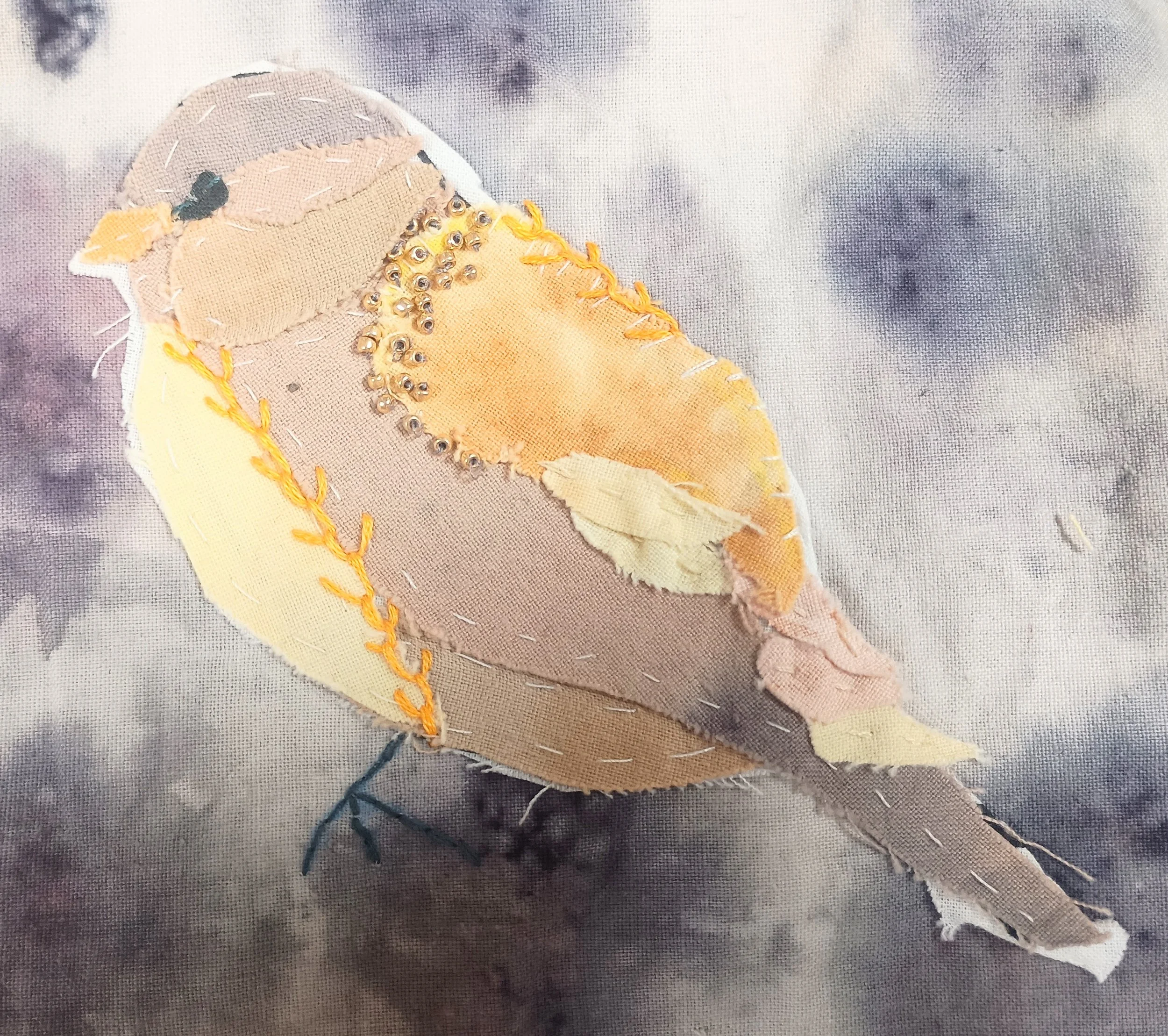 A fabric applique of a yellow bird with detailed embellishments, sewn onto a textured background.