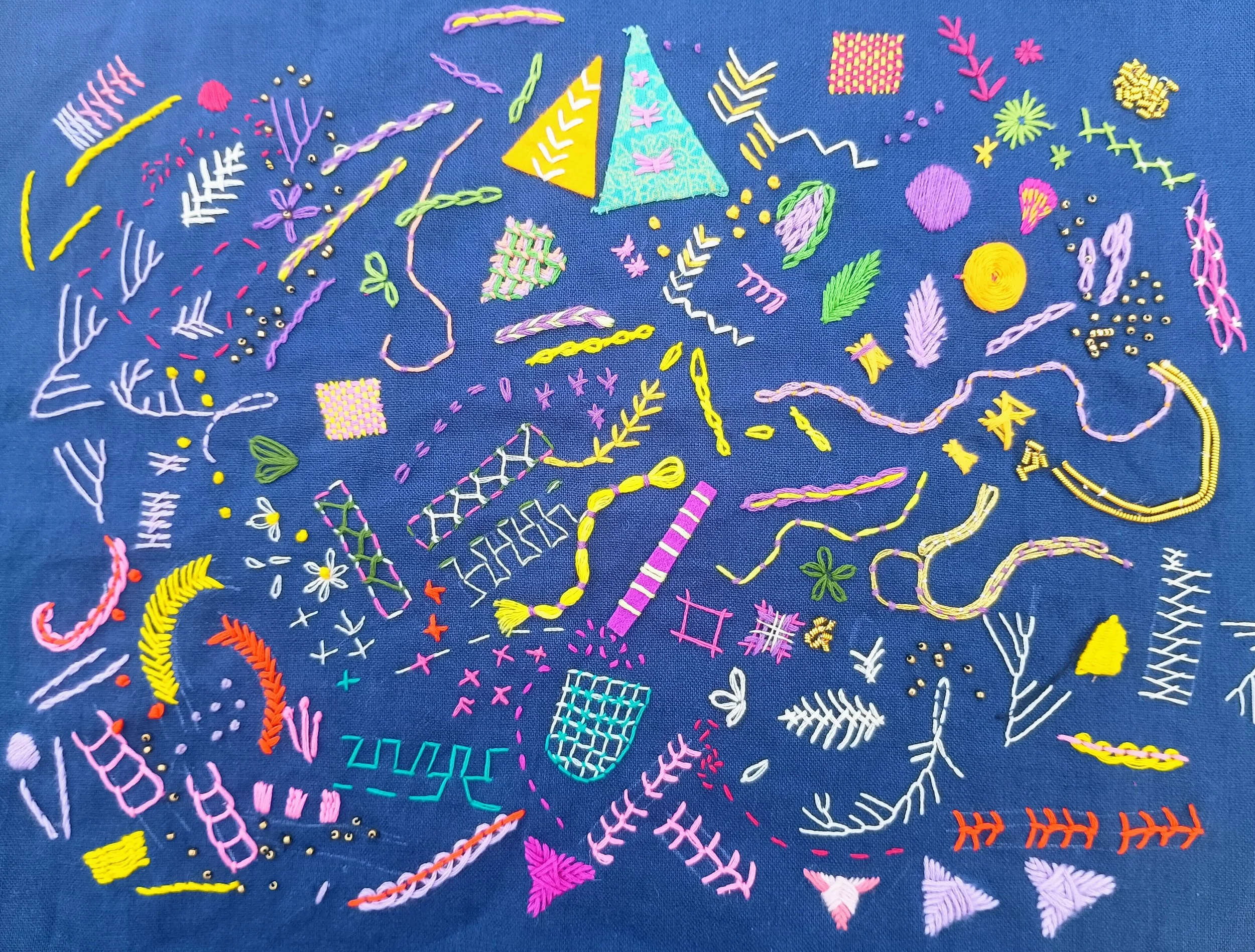 Colorful embroidery on a blue fabric background featuring various geometric shapes, lines, leaves, flowers, and abstract designs in pink, yellow, green, purple, and white.