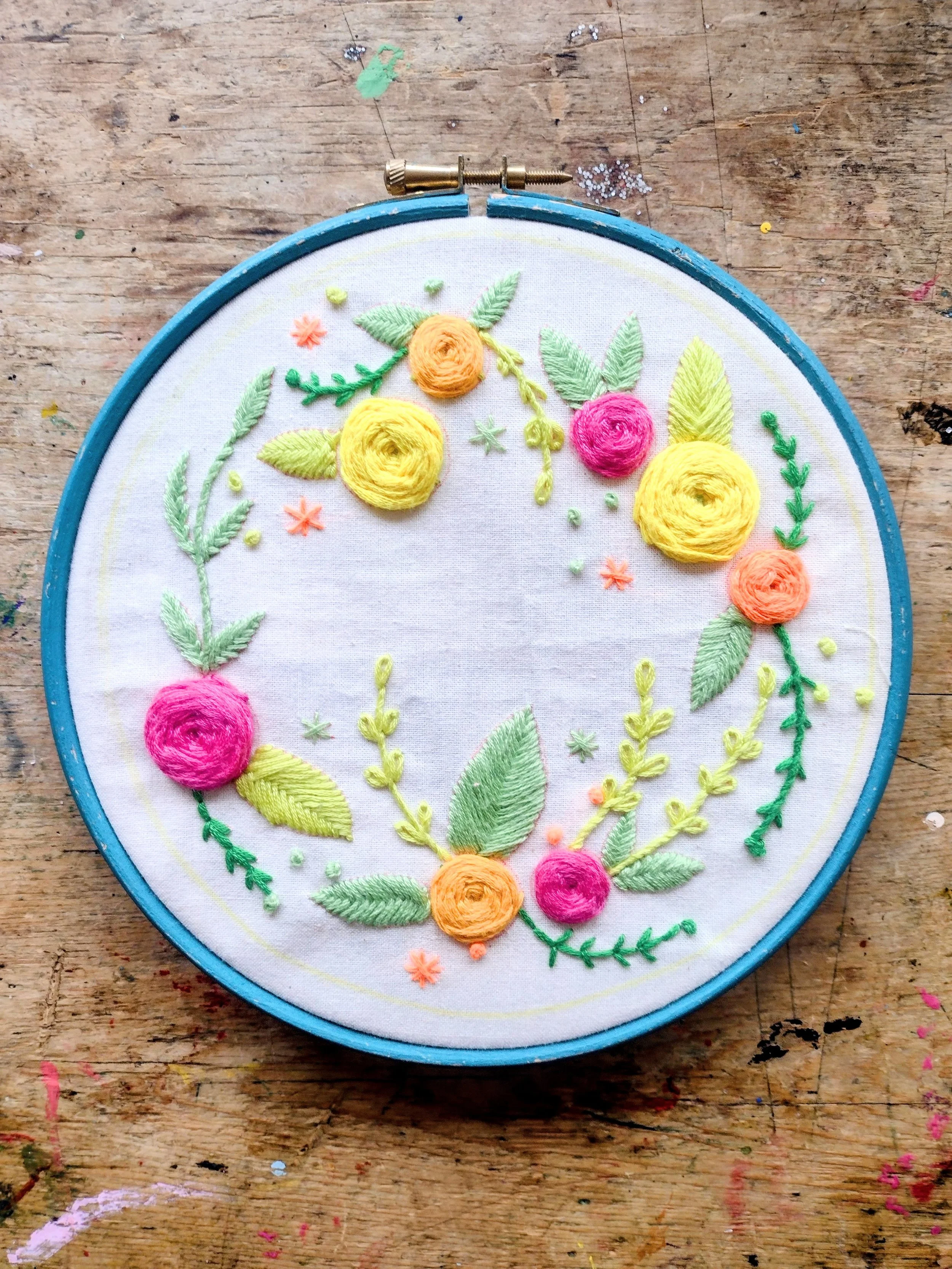 Embroidery hoop with colorful floral embroidery on white fabric, placed on a wooden surface with paint and craft supplies