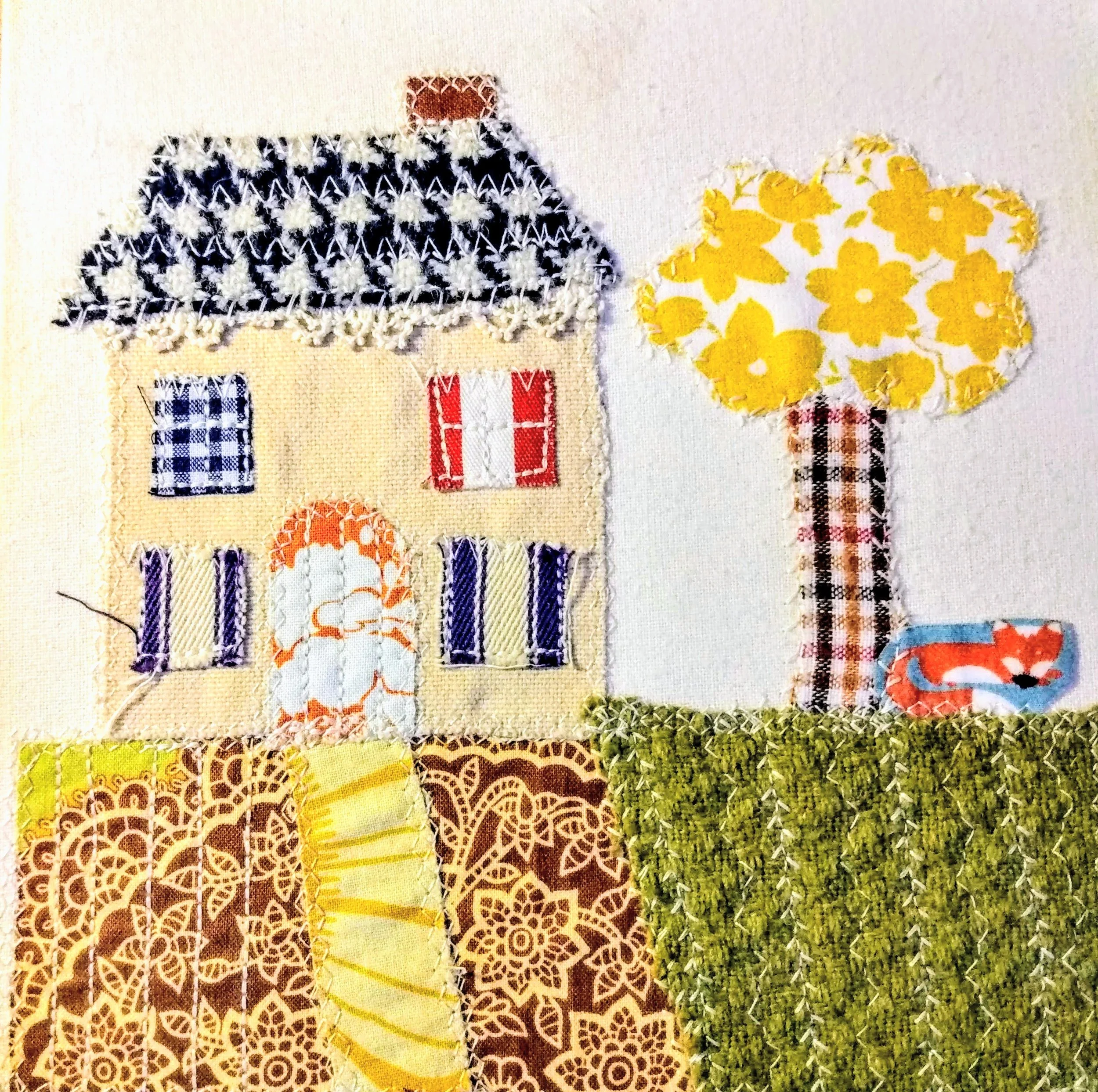 Close-up view of a textile art piece depicting a house with a black and white patterned roof, red and white door, blue and white checkered window, and surrounded by yellow, brown, and green patterned fabric or patches.