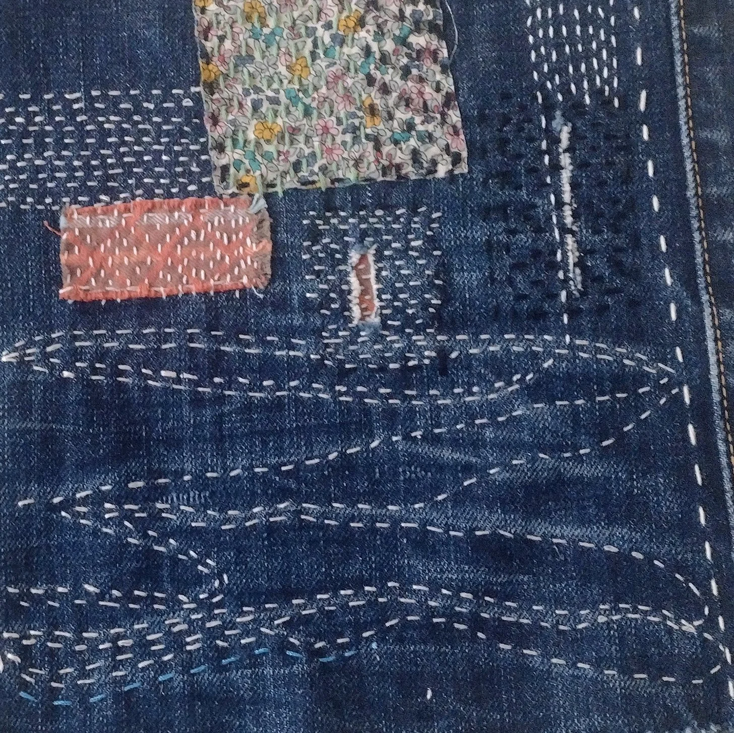 Close-up of a dark blue denim fabric with decorative white stitching forming patterns. Three small fabric patches with different floral and abstract designs are sewn onto the fabric.