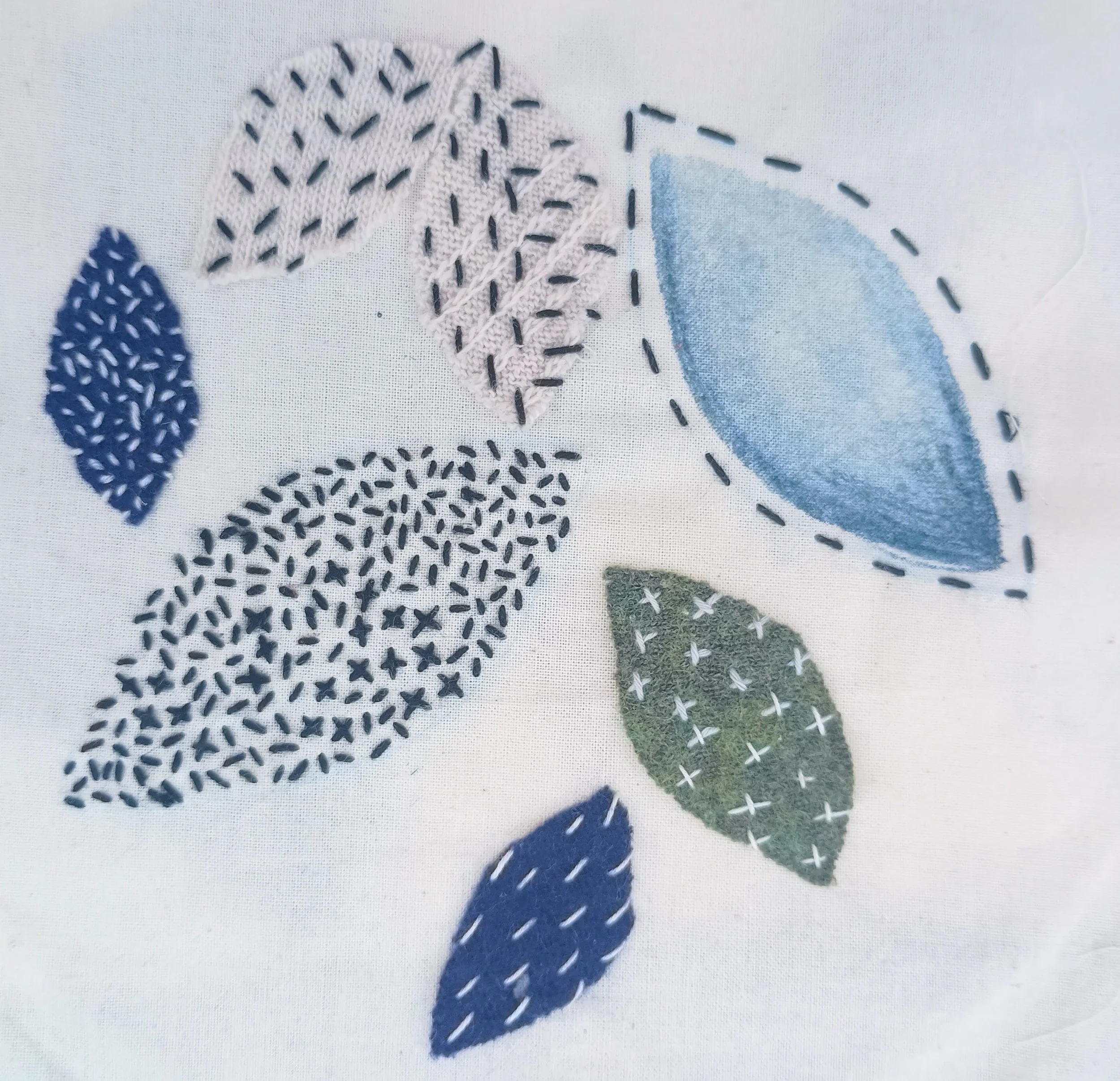 Close-up of embroidered leaves in blue, white, green, and purple on fabric, with visible stitch details.