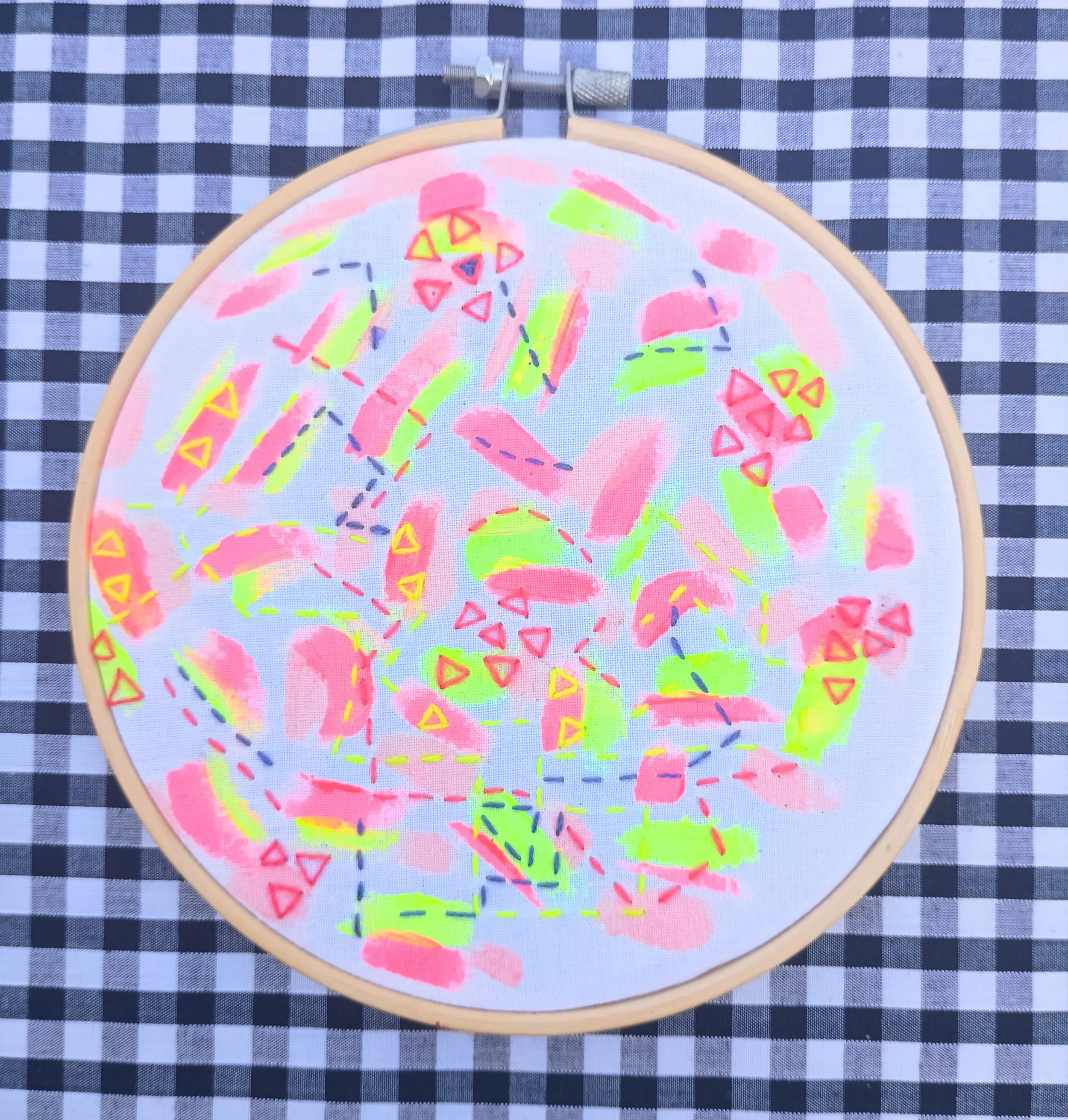 Embroidery hoop with a partially visible colorful abstract design on fabric, placed on a black and white checkered surface.