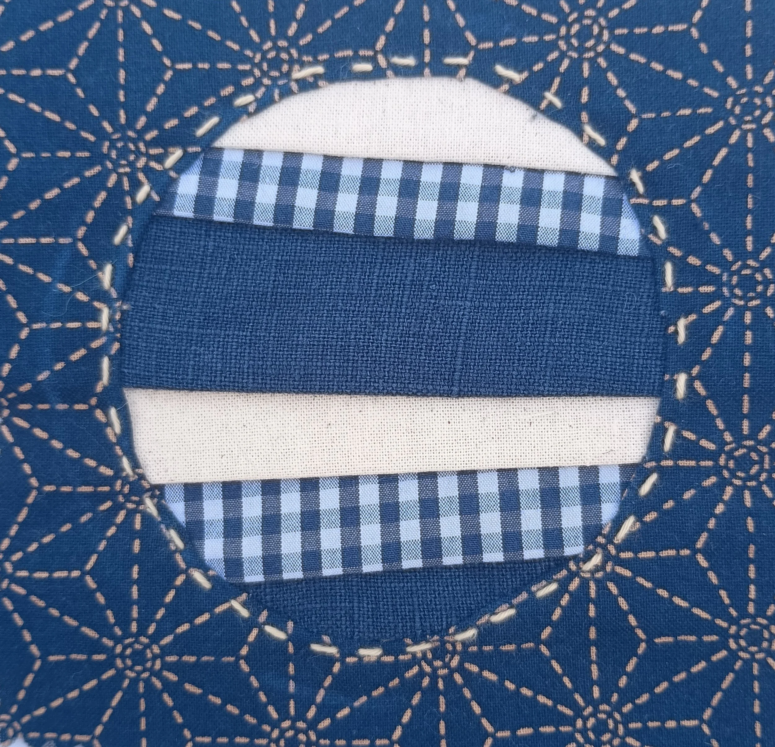 Close-up of a fabric with a navy blue background and pink dashed embroidered lines forming geometric patterns. Part of a white and blue checkered cloth is visible underneath, with a white fabric backing.